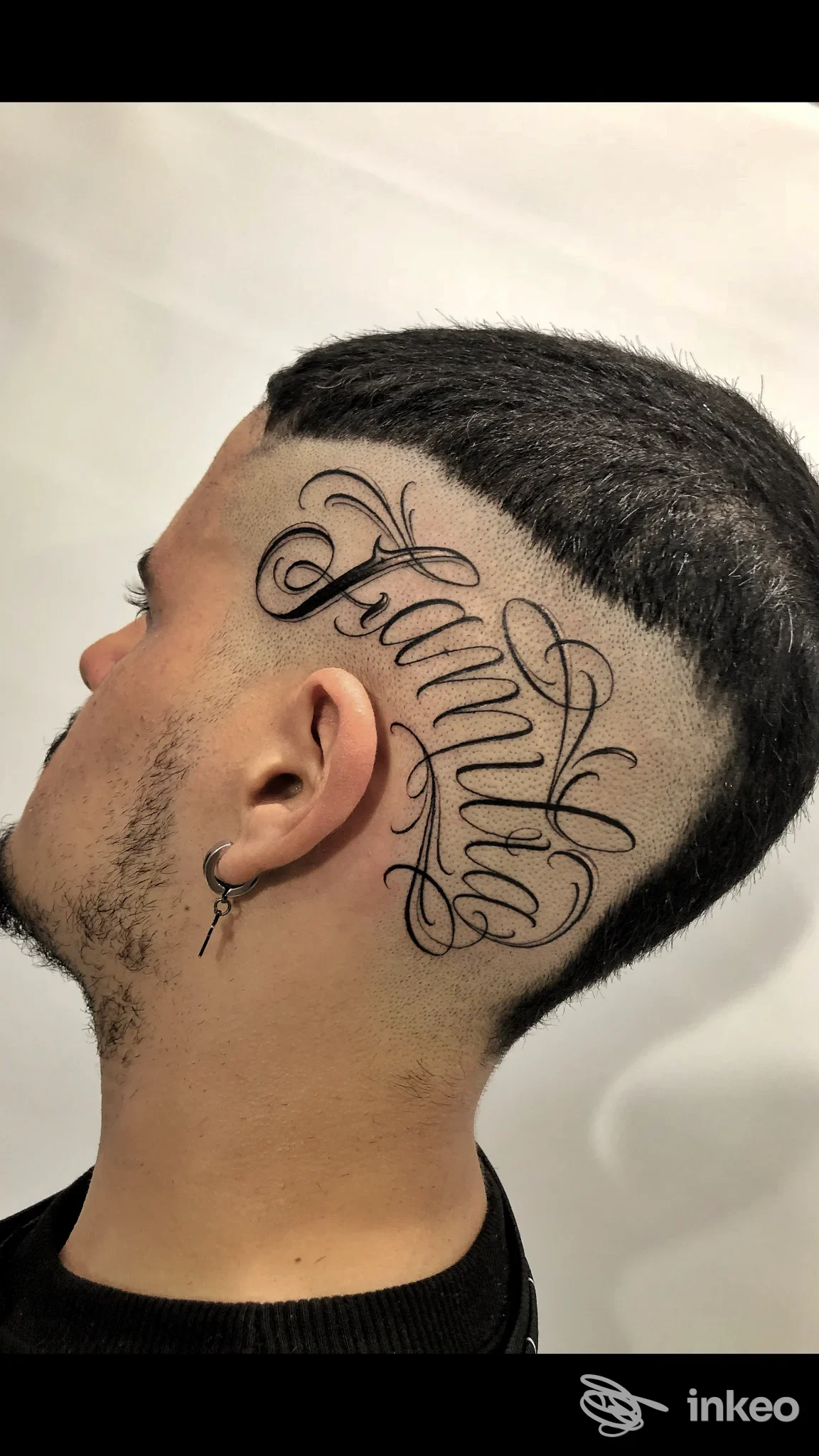 Parte laterale della testa con tatuaggio script elegante in nero, visibile dietro l’orecchio e sulla tempia, in primo piano su sfondo chiaro.