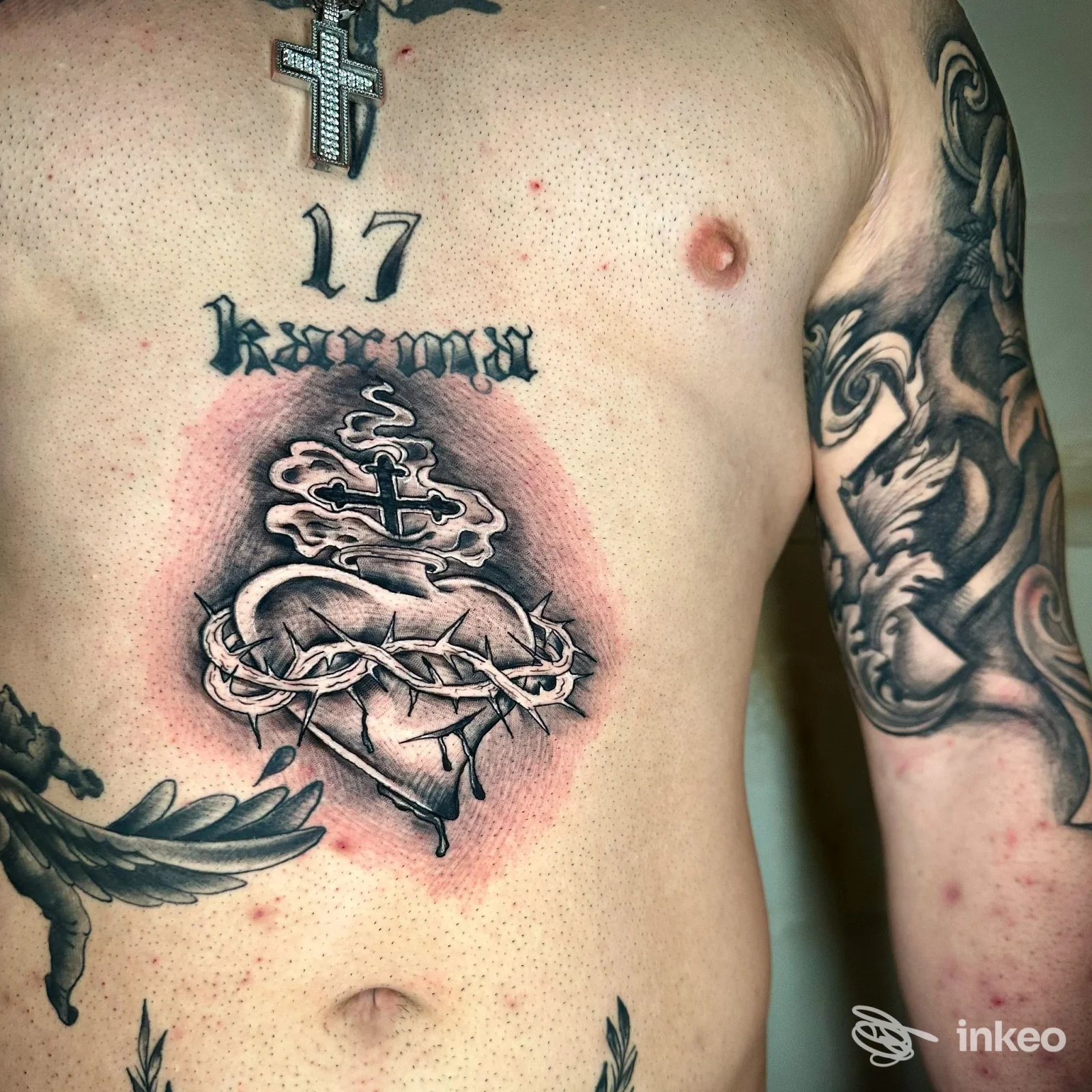 Tatuaggio sul torace raffigurante cuore con corona di spine e croce, sfondo rosso attorno al disegno, visibile sulla pelle maschile.