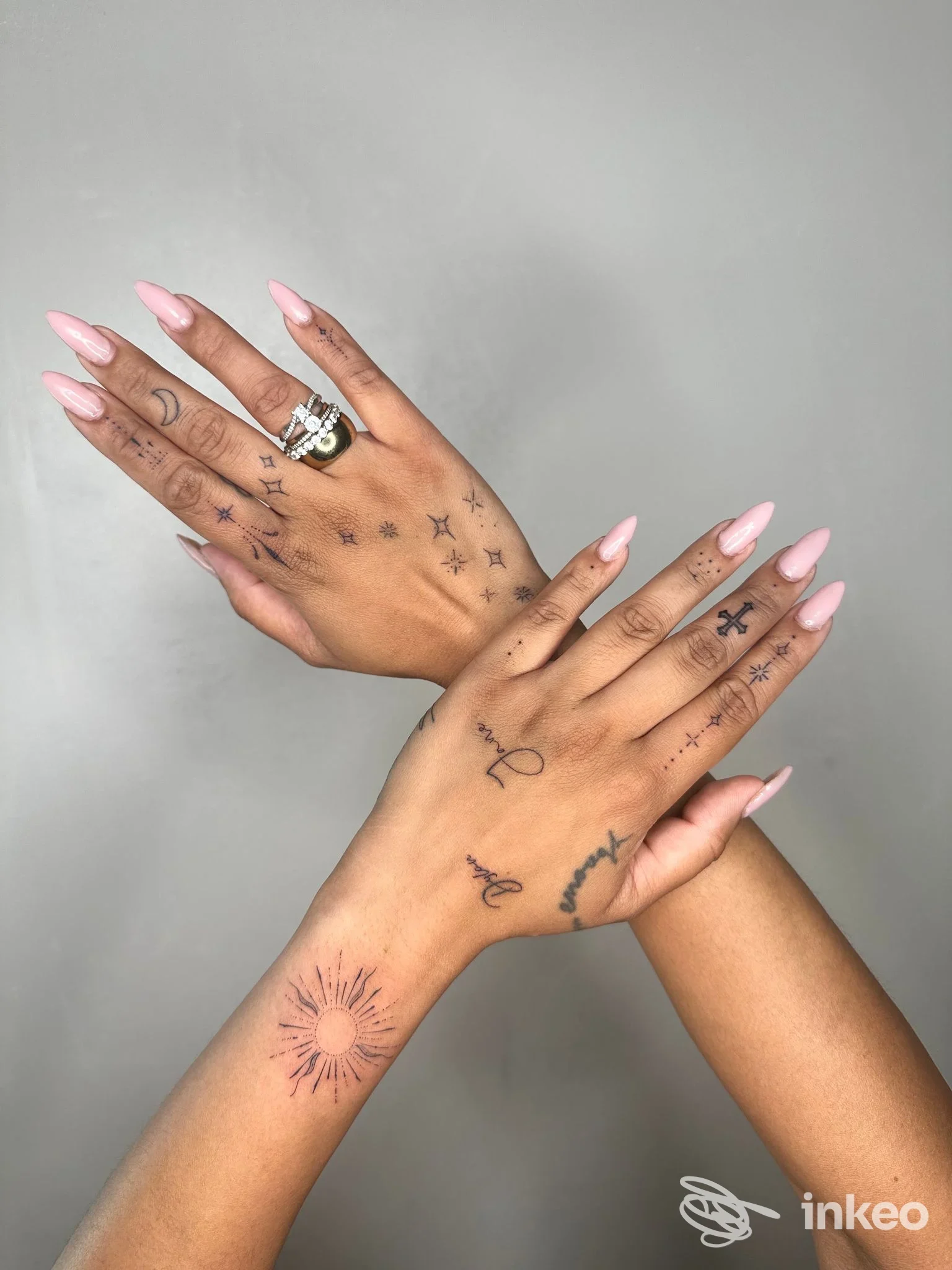 Due mani con tatuaggi su dita e dorso, stelle sul dorso e decorazioni sulle dita; unghie a mandorla rosa chiaro, sfondo uniforme. Firma Monros Tattoo non visibile, città non specificata.