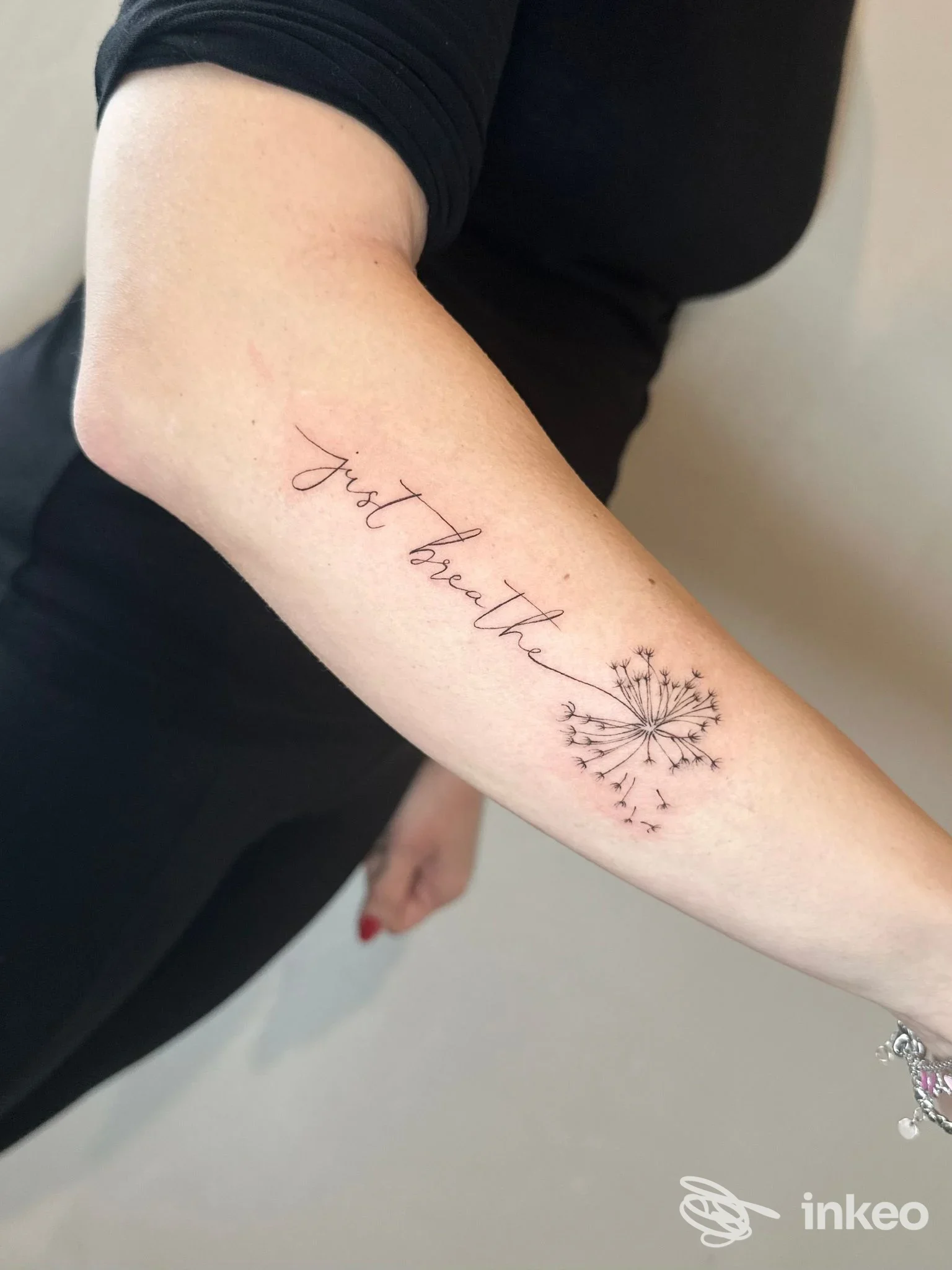 Tattoo sull’avambraccio sinistro: una scritta elegante in corsivo che dice "Just breath" con un disegno stilizzato di fiore/dente di leone che emana semi.