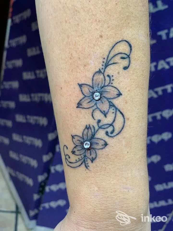 Tatuaggio floreale su pelle: due fiori stilizzati collegati da una linea curva, linea blu scuro con dettagli leggeri, tatuaggio visibile sull’avambraccio. studio Bull Tattoo & Body Piercing Studio, Napoli.