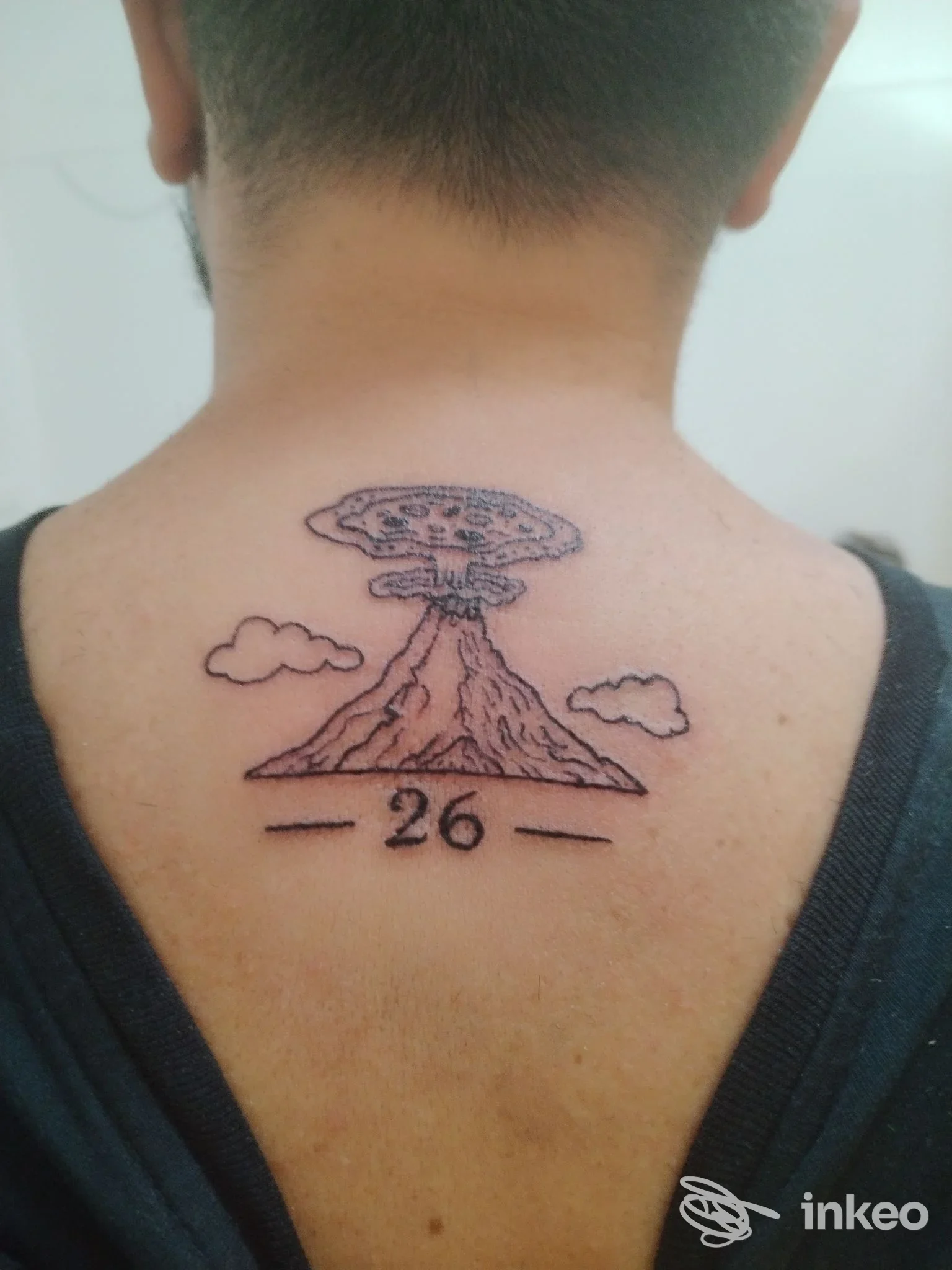 Tattoo sulla schiena di una persona: un vulcano in eruzione con nuvole ai lati e il numero 26 inscritto sotto, disegno in linee nere.