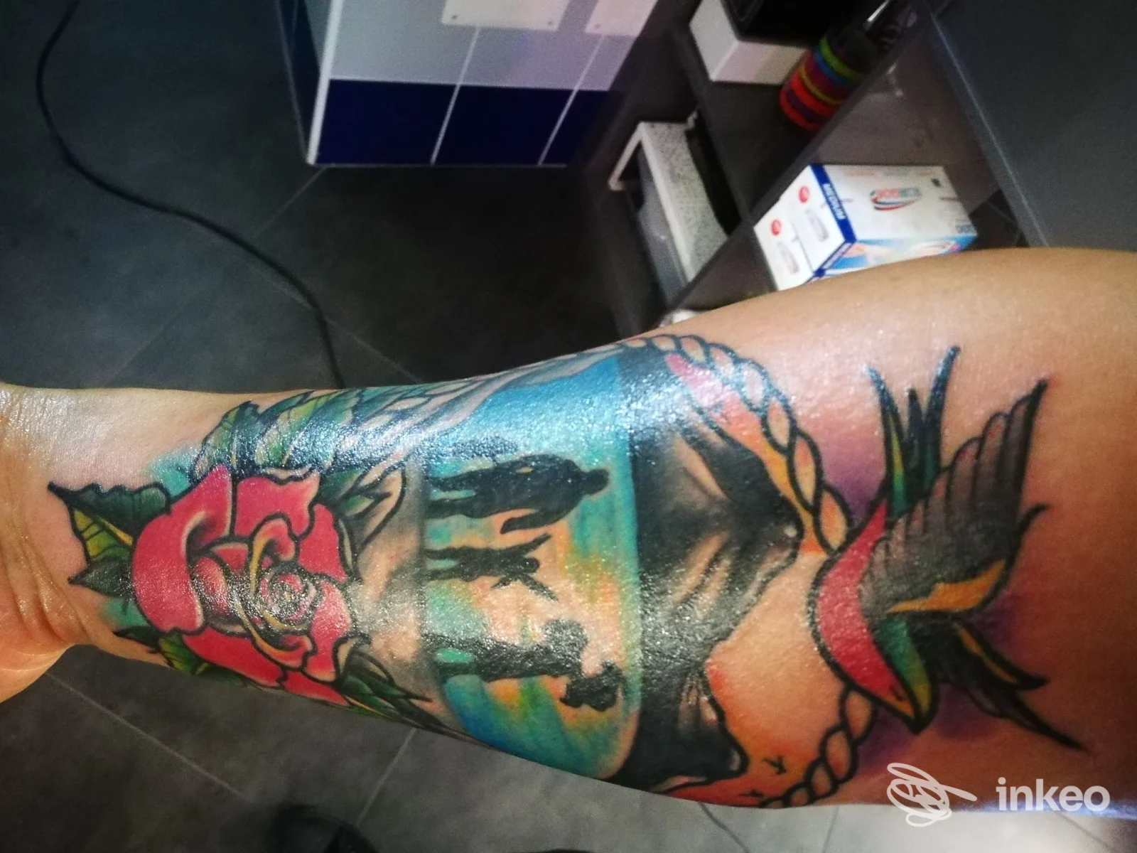 Braccio con tatuaggio a colori di Oplà Art Tattoo & Piercing, Castellammare di Stabia: rosa rossa, paesaggio marino e elementi intrecciati ben definiti e vividi.
