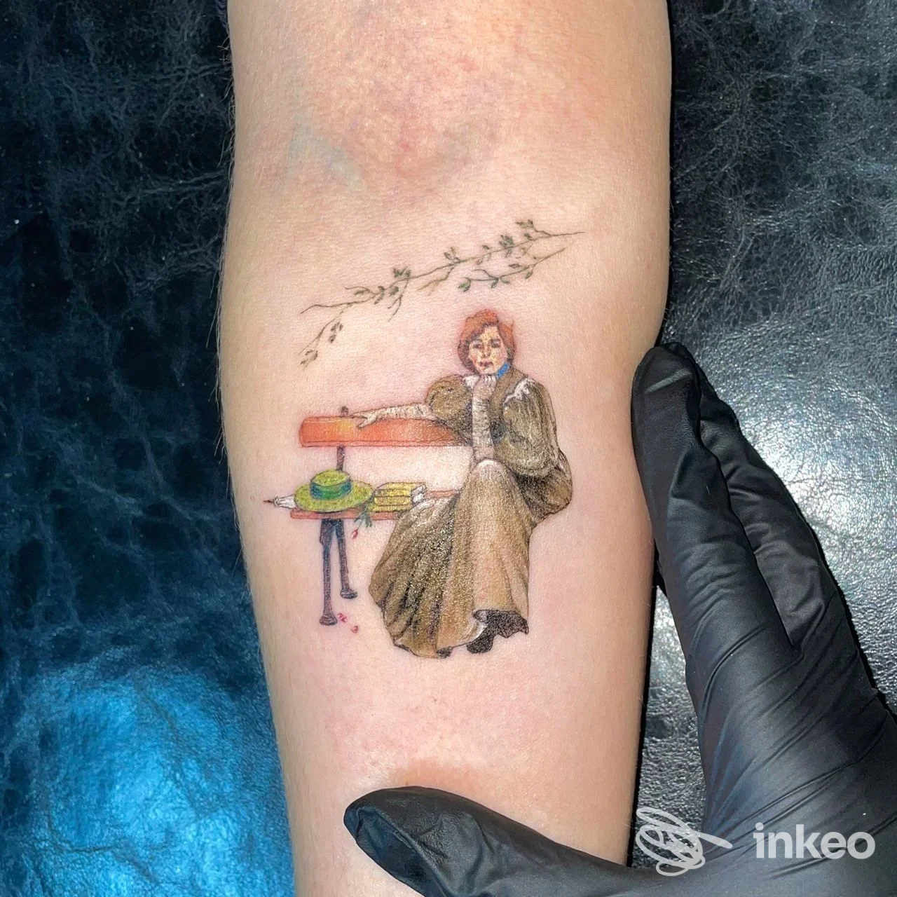 Tatuaggio sull'avambraccio raffigurante una persona seduta con un lungo oggetto arancione, decorazioni floreali tra i rami, realizzazione visibile sulla pelle, stile realistico.