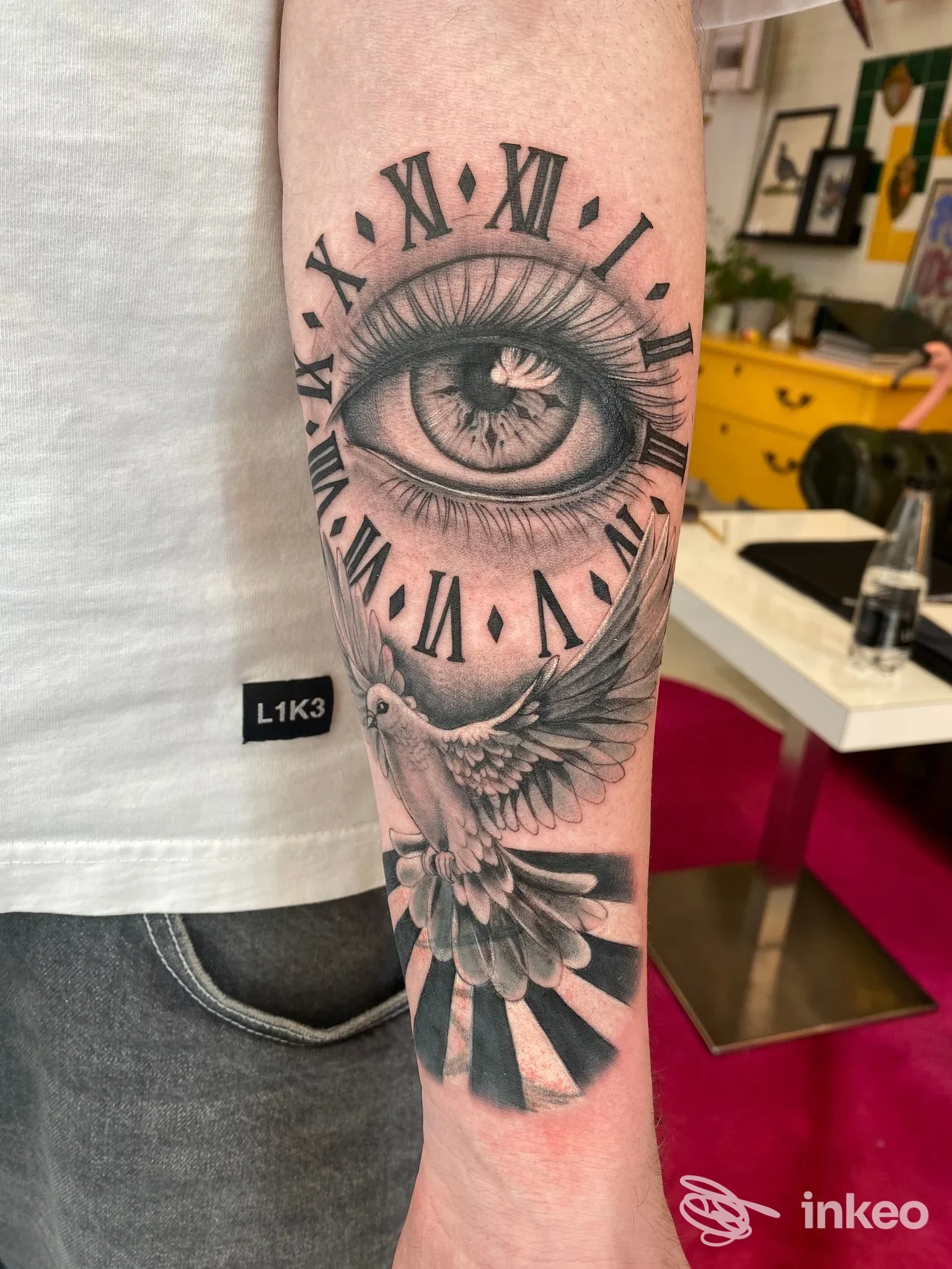 Tattoo sull’avambraccio raffigurante un grande occhio circondato da un orologio con numeri romani, con ali stilizzate che si estendono dal polpaccio verso la parte inferiore dell’avambraccio.