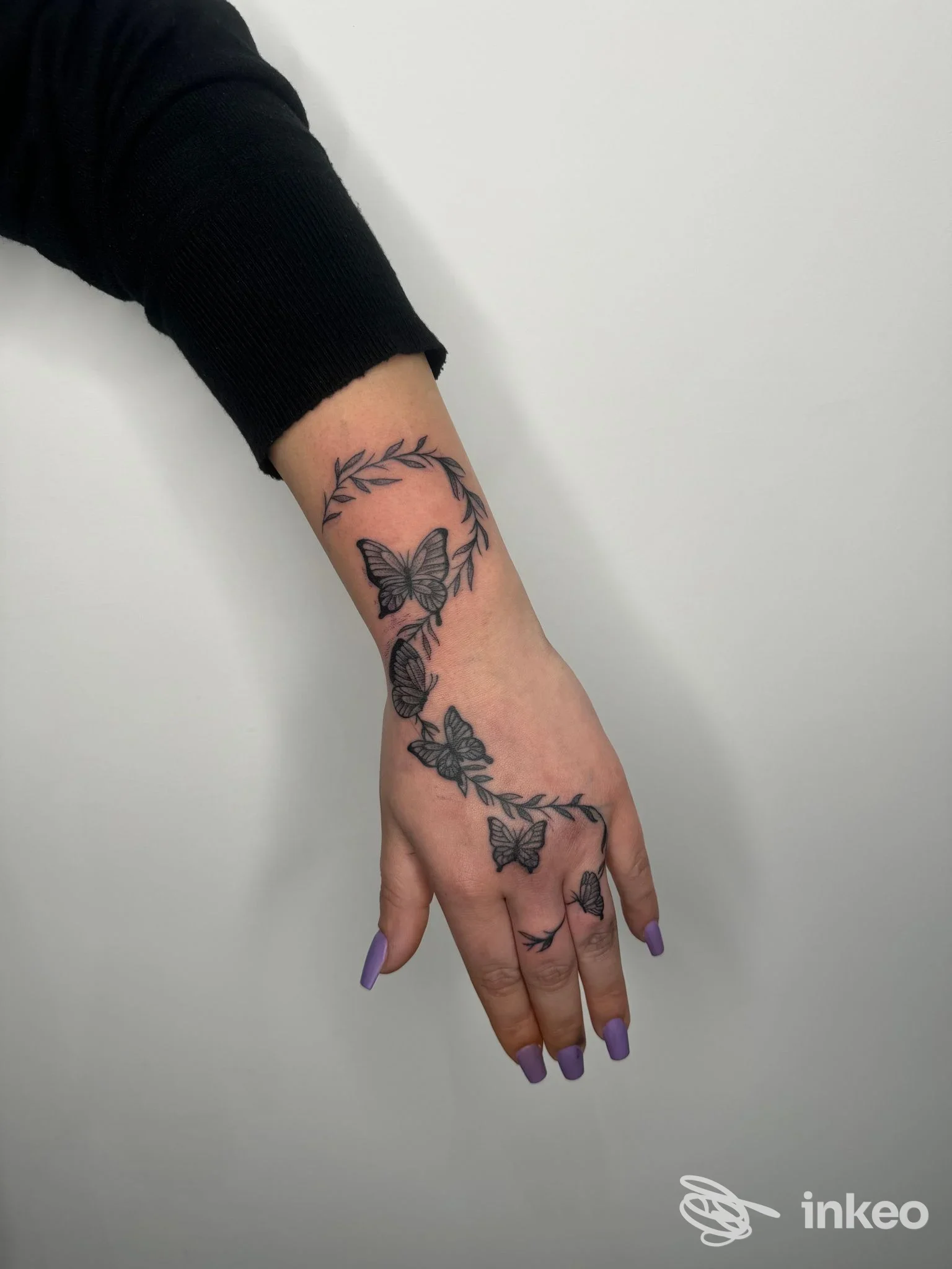 Mano sinistra con tatto floreale a farfalle lungo il dorso e le dita, tonalità nero-grigio, unghie viola, sfondo neutro.