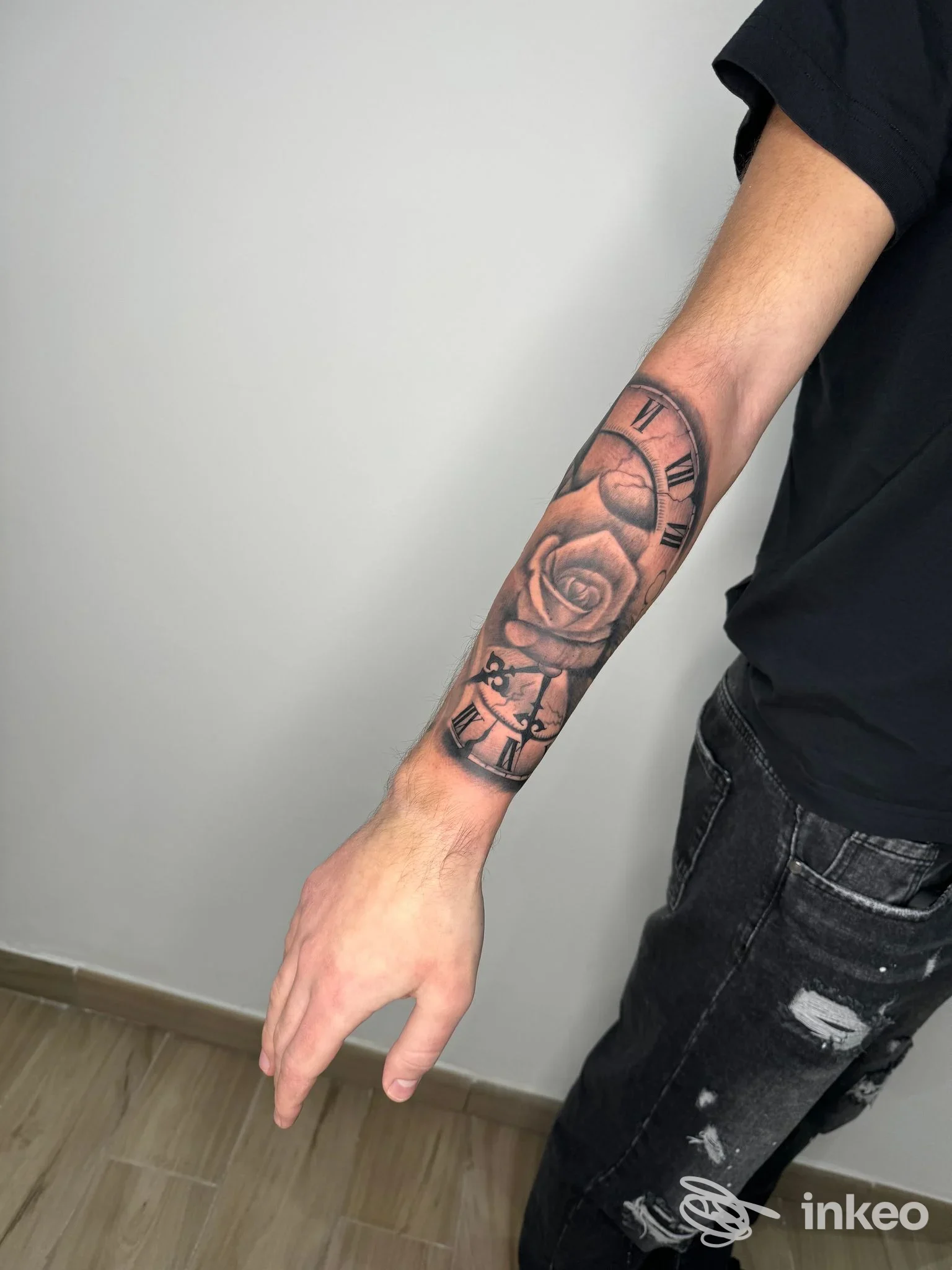 Braccio sinistro di un uomo con tatuaggio realistico in bianco e nero dalla spalla al polso: orologio romano, rosa e detalI floreali lungo tutta l'avambraccio; pelle visibile e contorni netti.