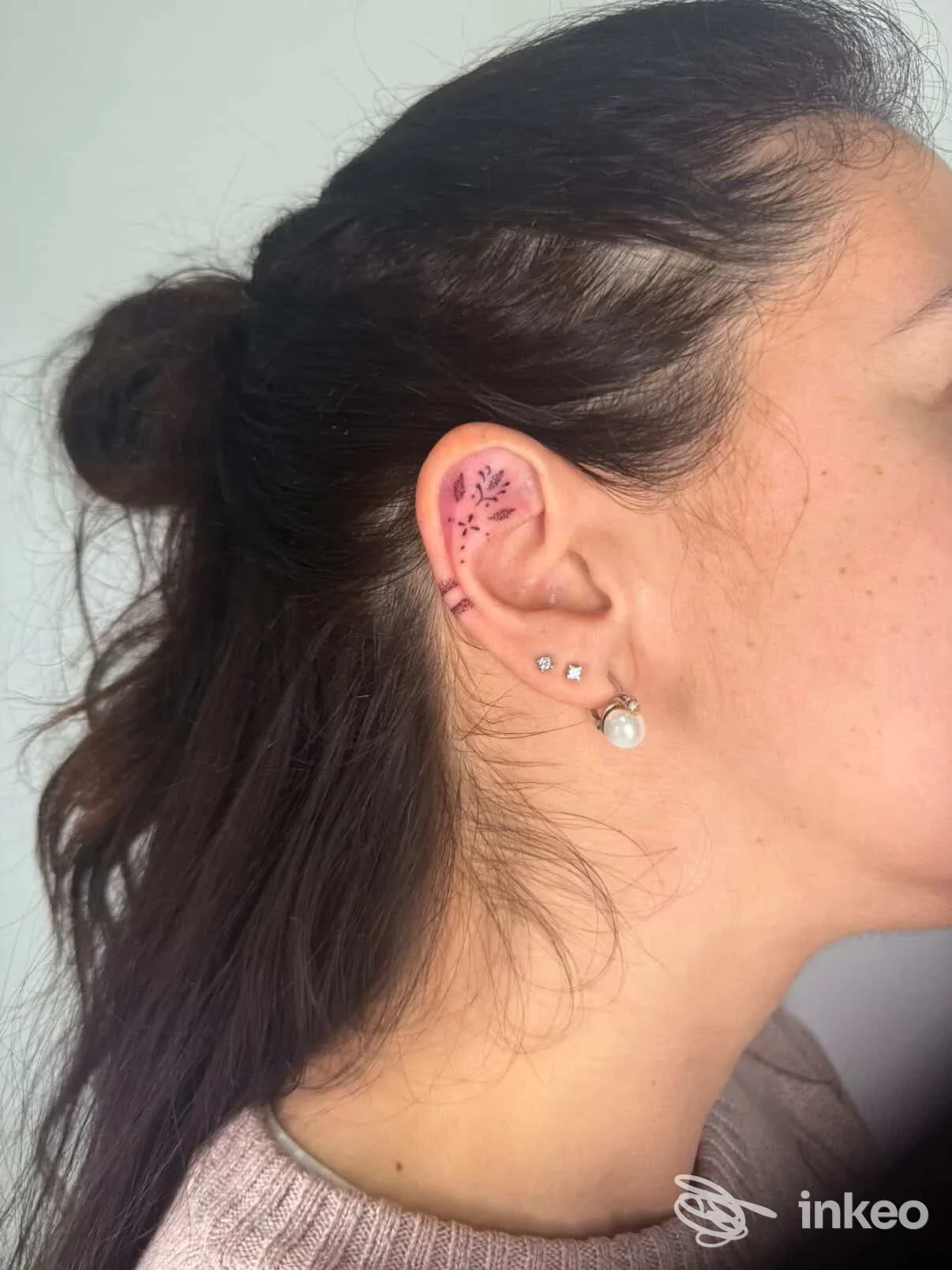 Orecchio di una persona con un piccolo tattoo floreale visibile sulla cartilage dell'orecchio, accompagnato da orecchini; in primo piano, senza mostrare il volto intero.