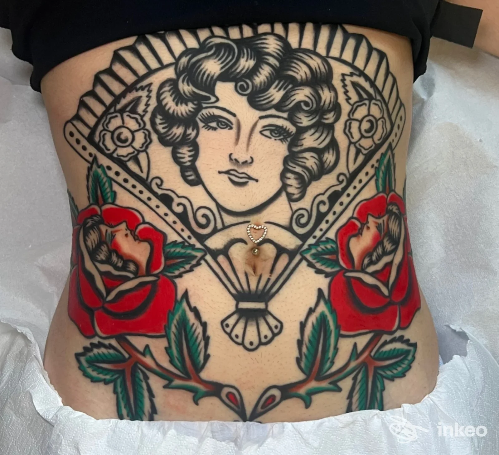 Tattoo tradizionale su pancia/fianco: figura femminile con capelli ricci, cornice con rose rosse e foglie verdi, linee nere nette e colori vividi.