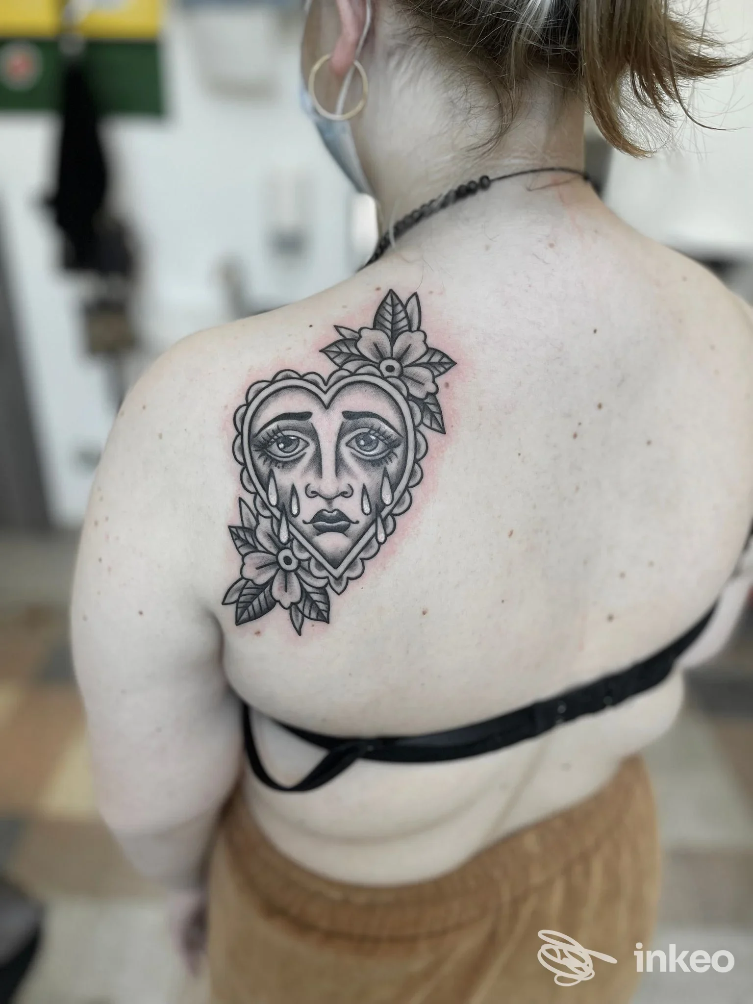 Tattoo sul dorso della spalla raffigurante un volto umano stilizzato all'interno di un cuore, con fiori intorno. Colori in bianco e nero, disegno lineare netto. Studio Wind Rose, Cava de' Tirreni.