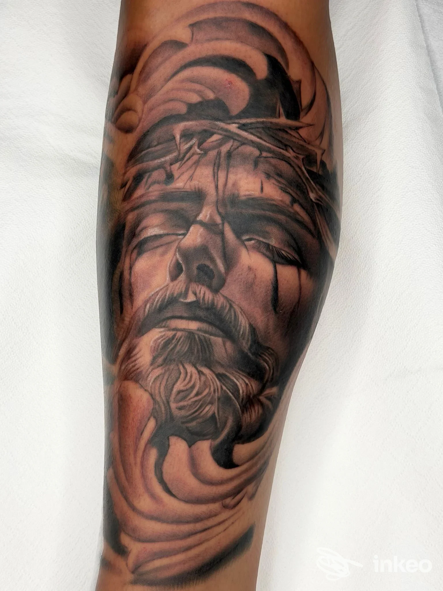 Tattoo realistico su avambraccio raffigurante figura religiosa con linee nere e grigie che enfatizzano la testa e la corona di spine; illuminazione neutra su sfondo chiaro.