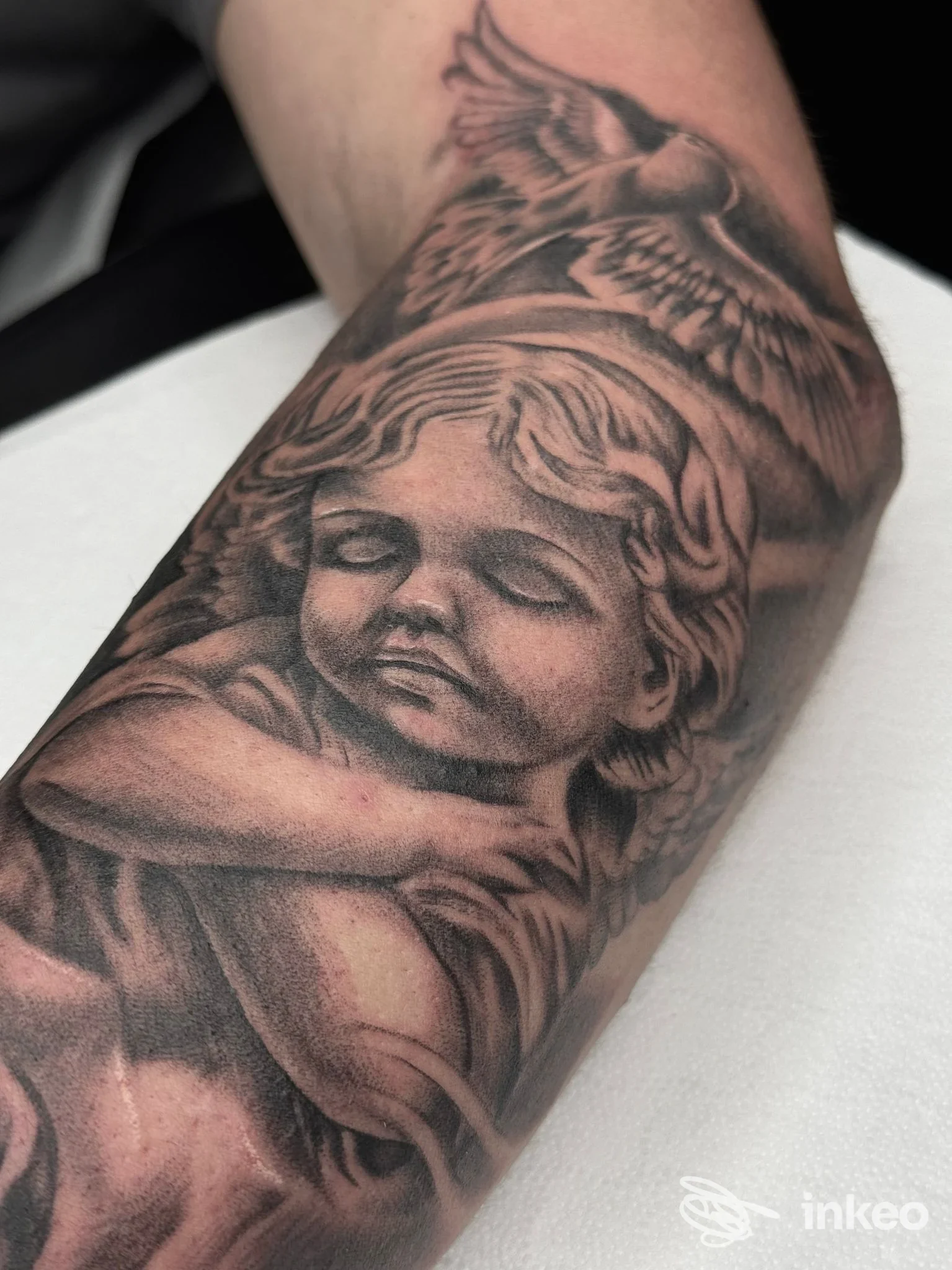 Tattoo realistico su avambraccio raffigurante un angelo bambino con ali, realizzato sulla pelle, senza elementi di sovrapposizione di social o schermi.