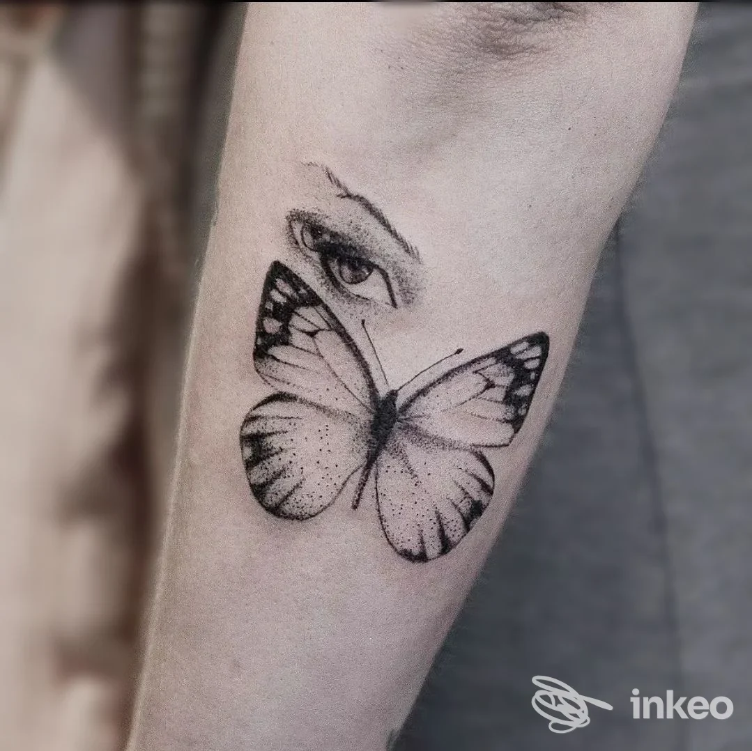 Tattoo sul braccio raffigurante una farfalla in bianco e nero, con un occhio parziale visibile sopra; realizzo corporeo chiaramente distinguibile.