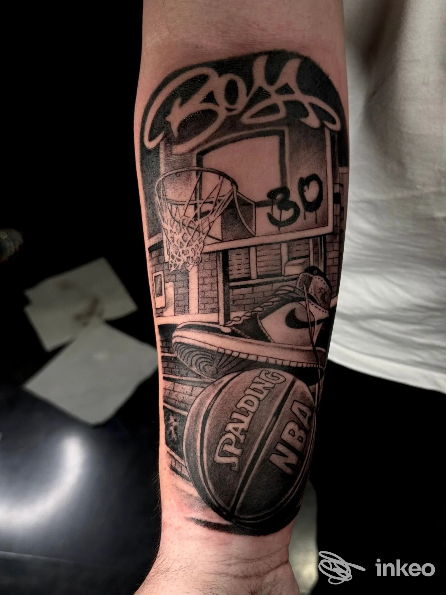 Tattoo realistico sul braccio raffigurante pallone NBA Slam Dunk e tabellone, in stile blackwork con sfondo scuro; disegno ben definito su pelle.