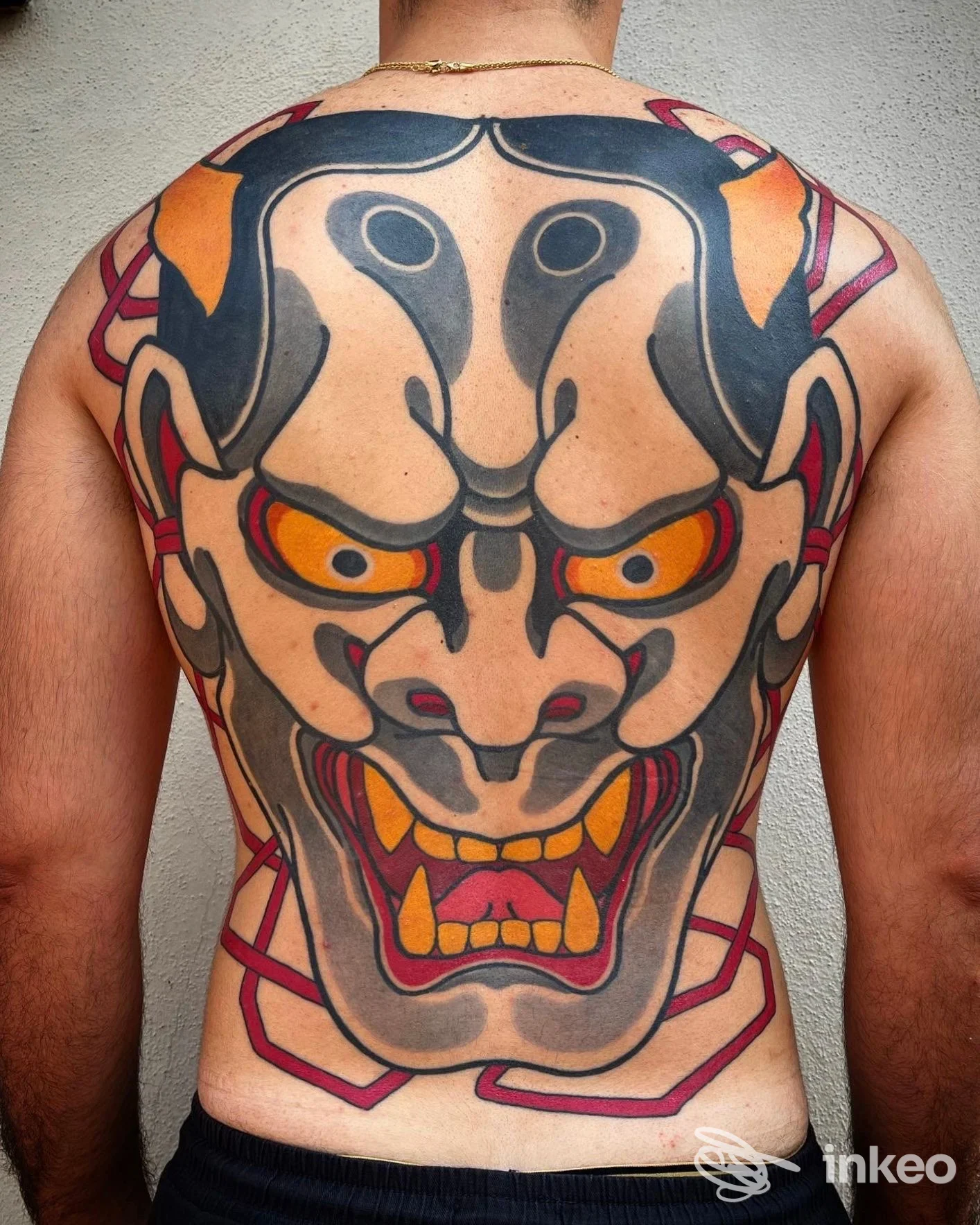 Schiena intera di un uomo con un grande tatuaggio tradizionale su volto demoniaco, colori nero, grigio, arancione e rosso, dal carattere orientale/pop contemporaneo.