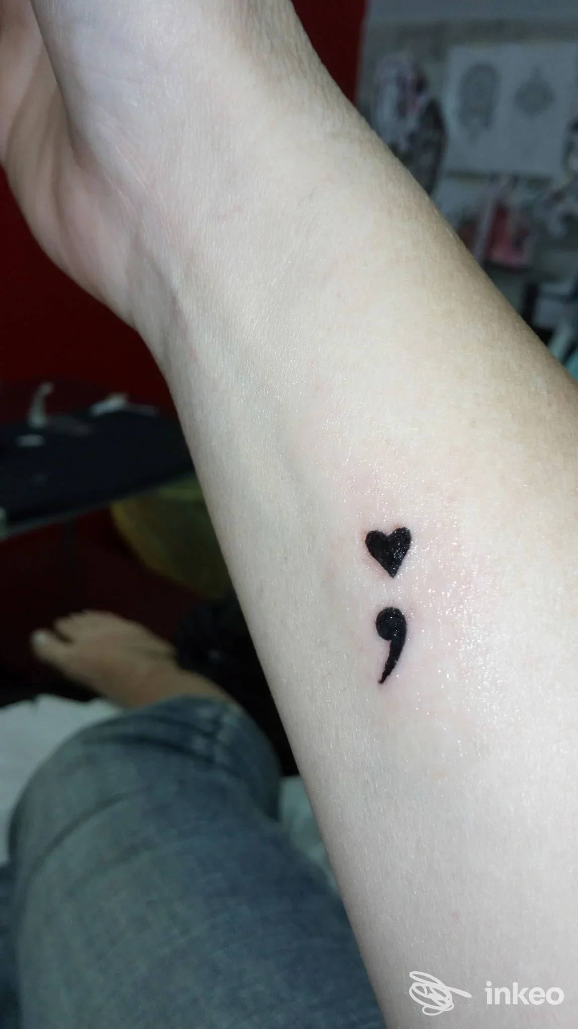 Piccolo tatuaggio visibile sull'avambraccio: cuore nero posto sopra un punto e virgola, senza presenza di volto o elementi social.