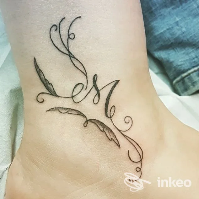 Tattoo grafico su caviglia con elementi floreali e lettere stilizzate, realizzato sulla pelle.