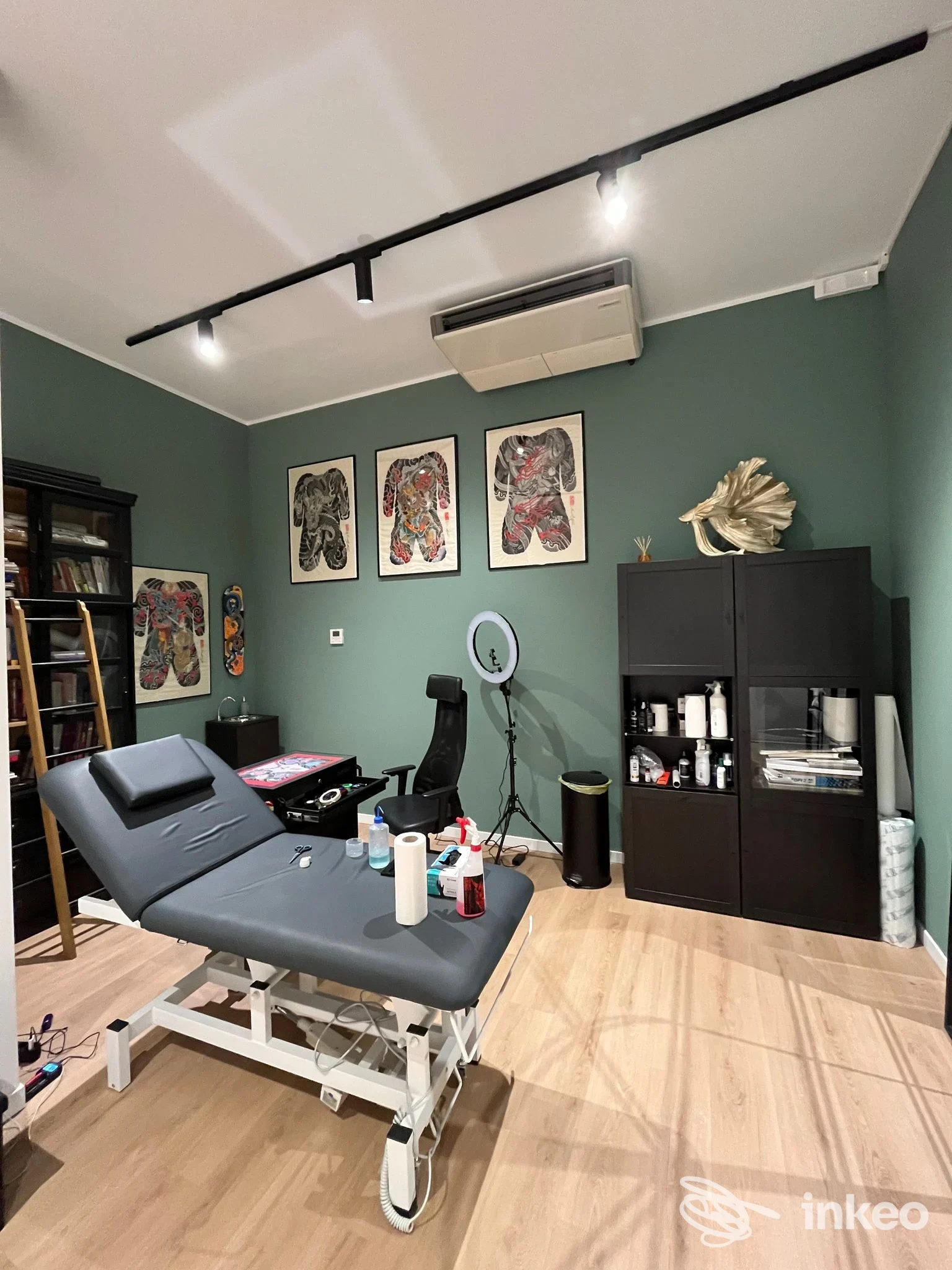 Interior di studio tatuaggi Punch Tattoo a Somma Lombardo: postazione con tavolo tatuaggio, sedie, scaffali e attrezzature, pareti verde scuro e decorazioni alle pareti.