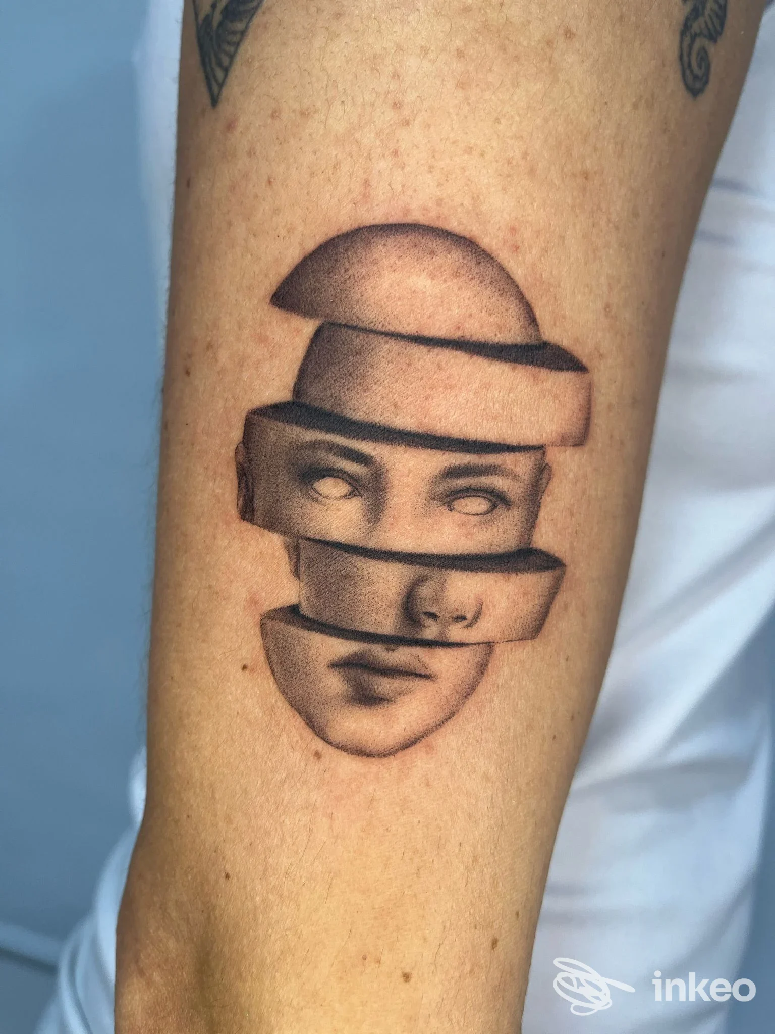 Tatuaggio realistico sul braccio raffigurante una faccia divisa in bande orizzontali, con dettagli di occhi e labbra parziali.