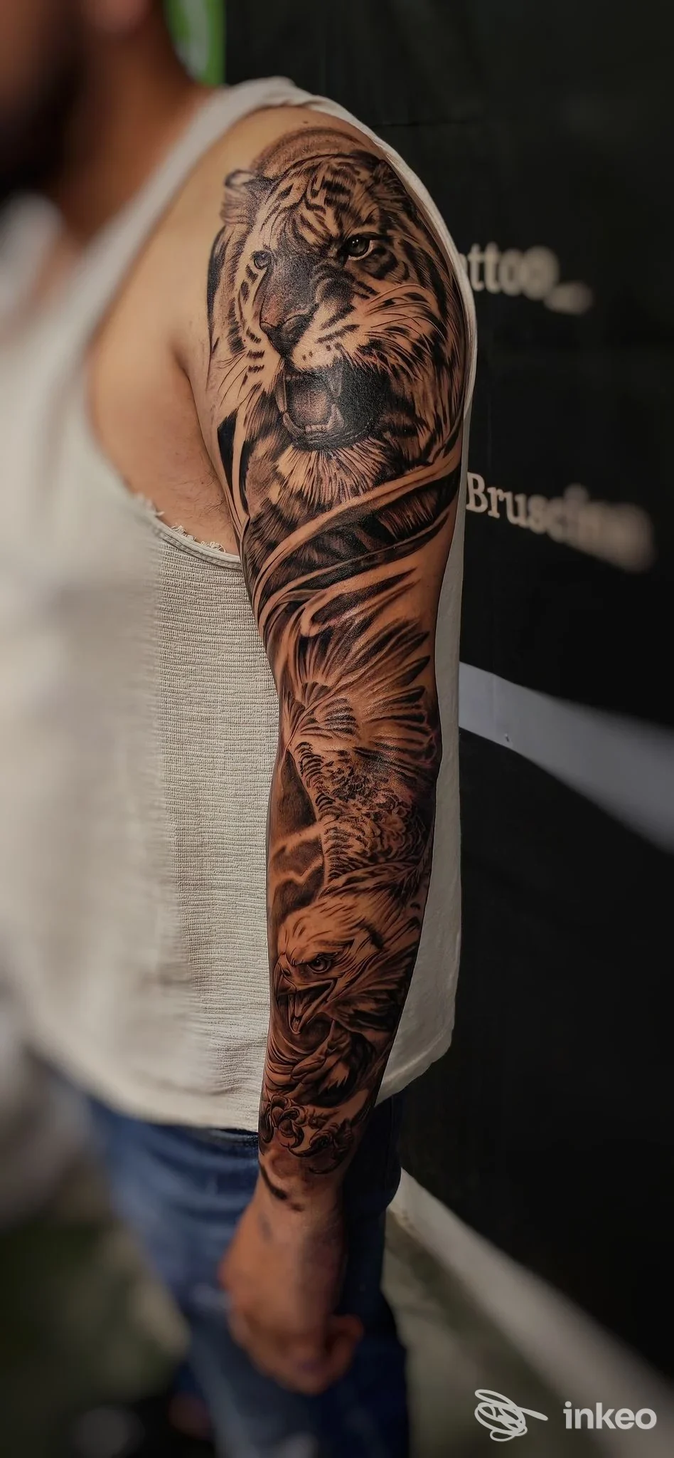 Tatuaggio realistico di leone sull'avambraccio, visibile su braccio di una persona con manica bianca; sfondo con bandiera nera.