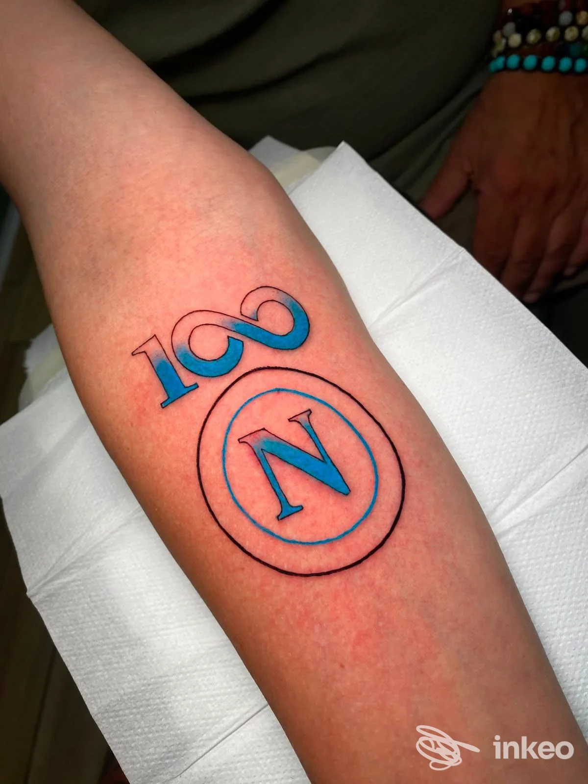 Tatuaggio originale su avambraccio: disegno lineare con cerchio contenente una lettera N, e numeri stilizzati blu-che rosso, su pelle chiara, eseguito presso Vitale Tattoo a Napoli.