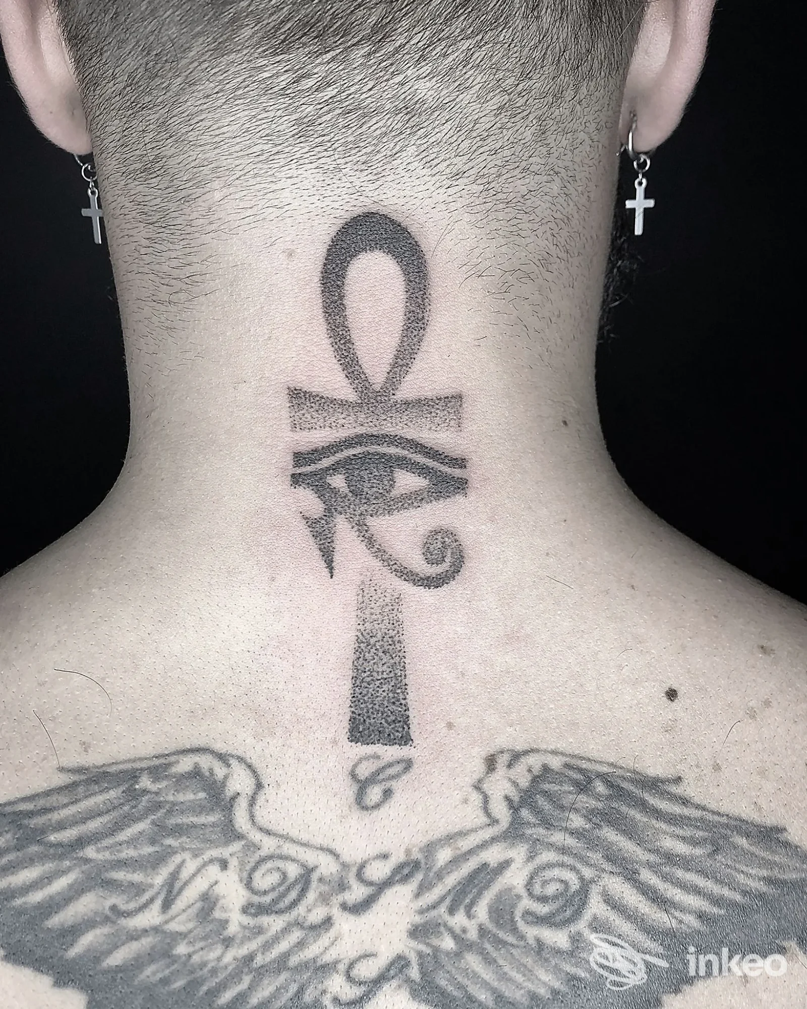 Tattoo visibile sulla nuca: un ank (croce con orecchio di Horus) sopra una figura del Taiw? simbolo, con ali tatuate sulla parte alta della schiena; stile inchiostro nero su pelle chiara, zona nuca.