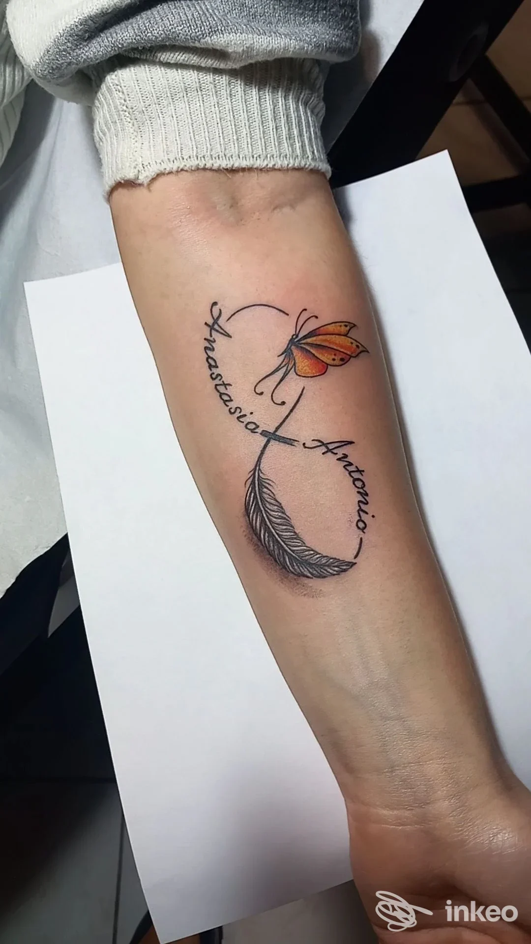 Avambraccio con tatuaggio di piuma e fiore stilizzato, testo elegante a forma di curva attorno al motivo.