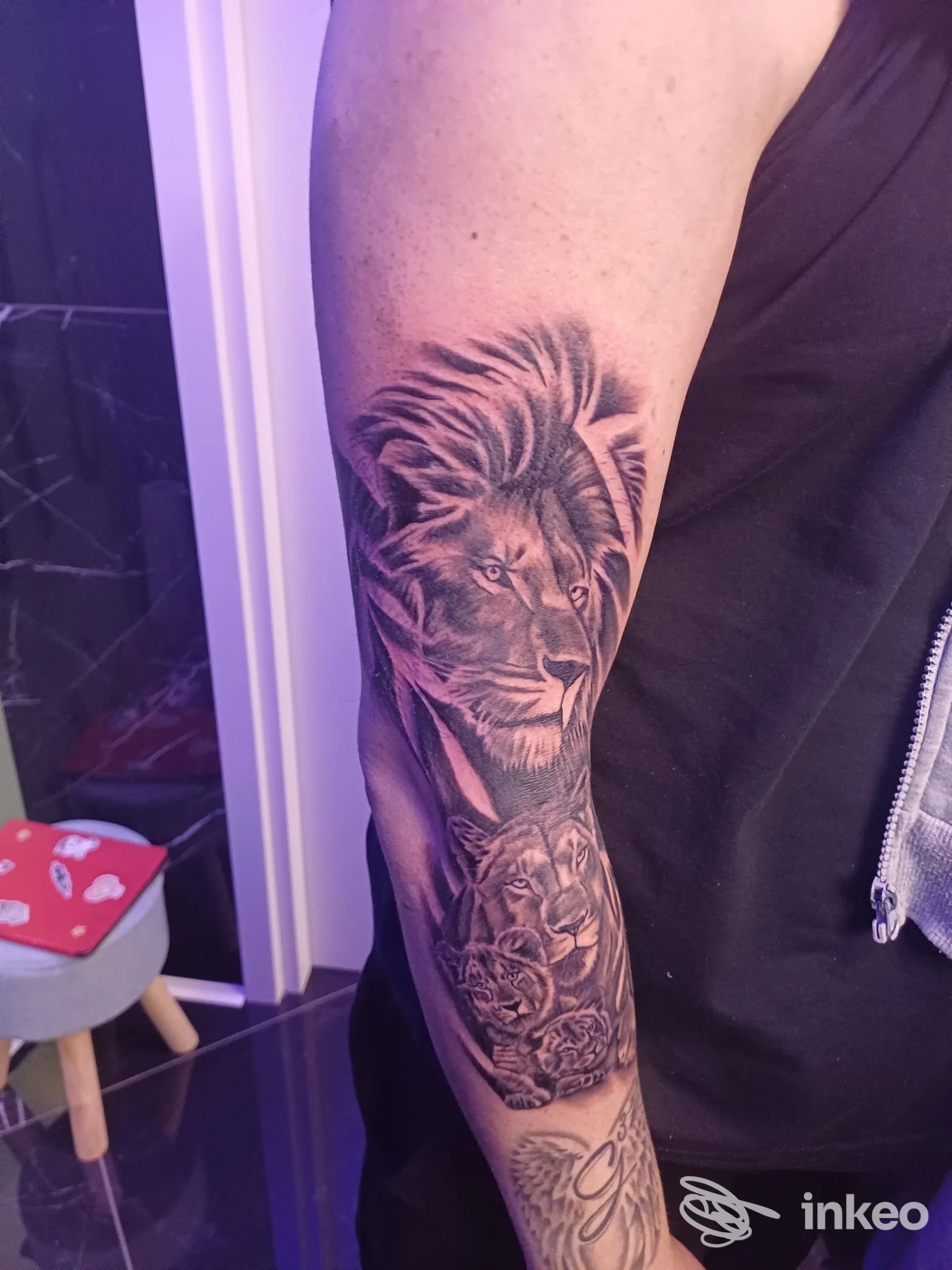 Braccio di una persona con un tatuaggio realistico a tema leone e un altro disegno sulla pelle, con tonalità bianco e nero, visibile in luce artificiale.