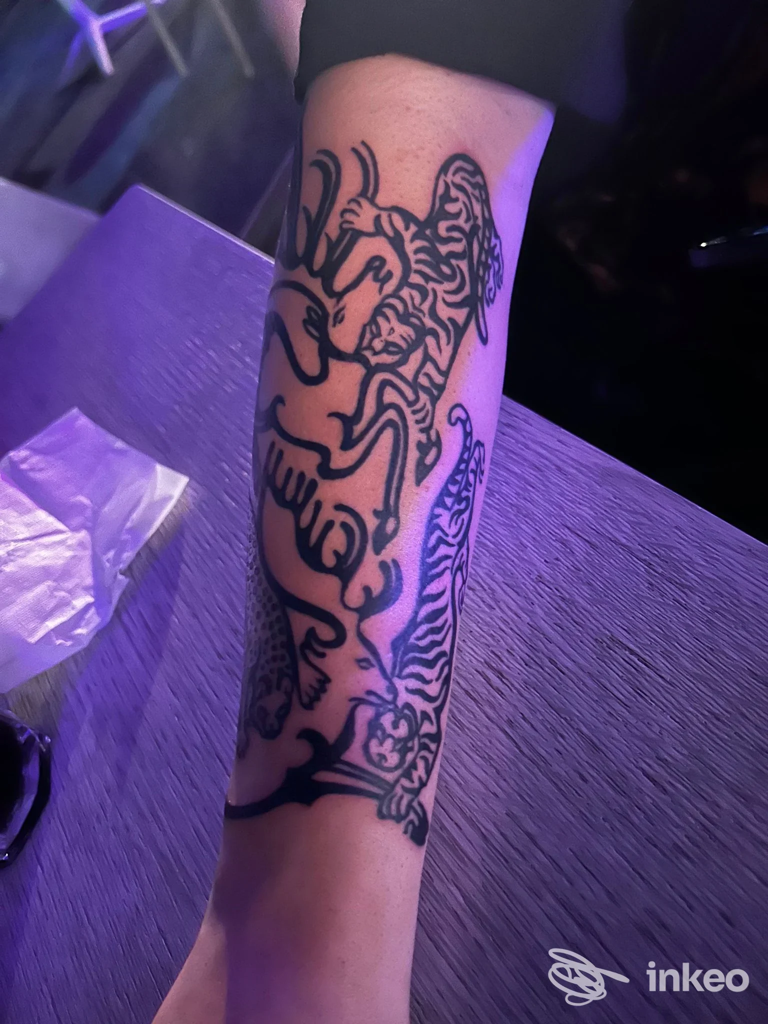Avambraccio visibile con tatuaggio in stile linework a linee nere, nessun volto visibile, illuminazione soffusa, sfondo tavolo scuro.