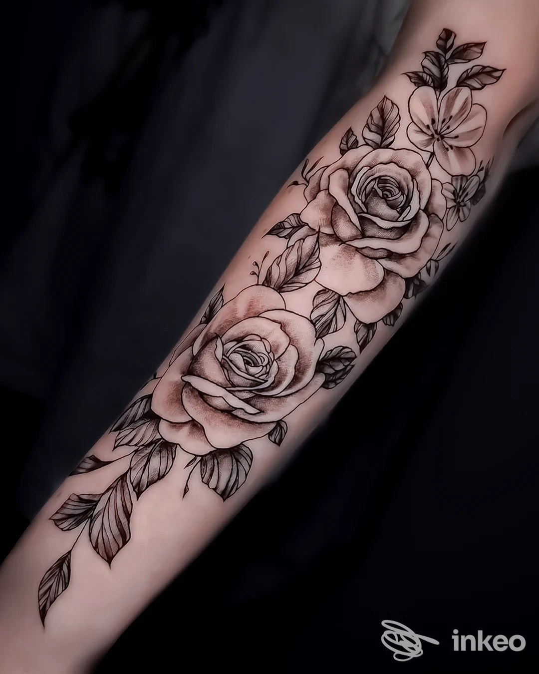 Tattoo sul braccio: significativo motivo di rose e fiori in bianco e nero su pelle chiara, scatto laterale con sfondo scuro.