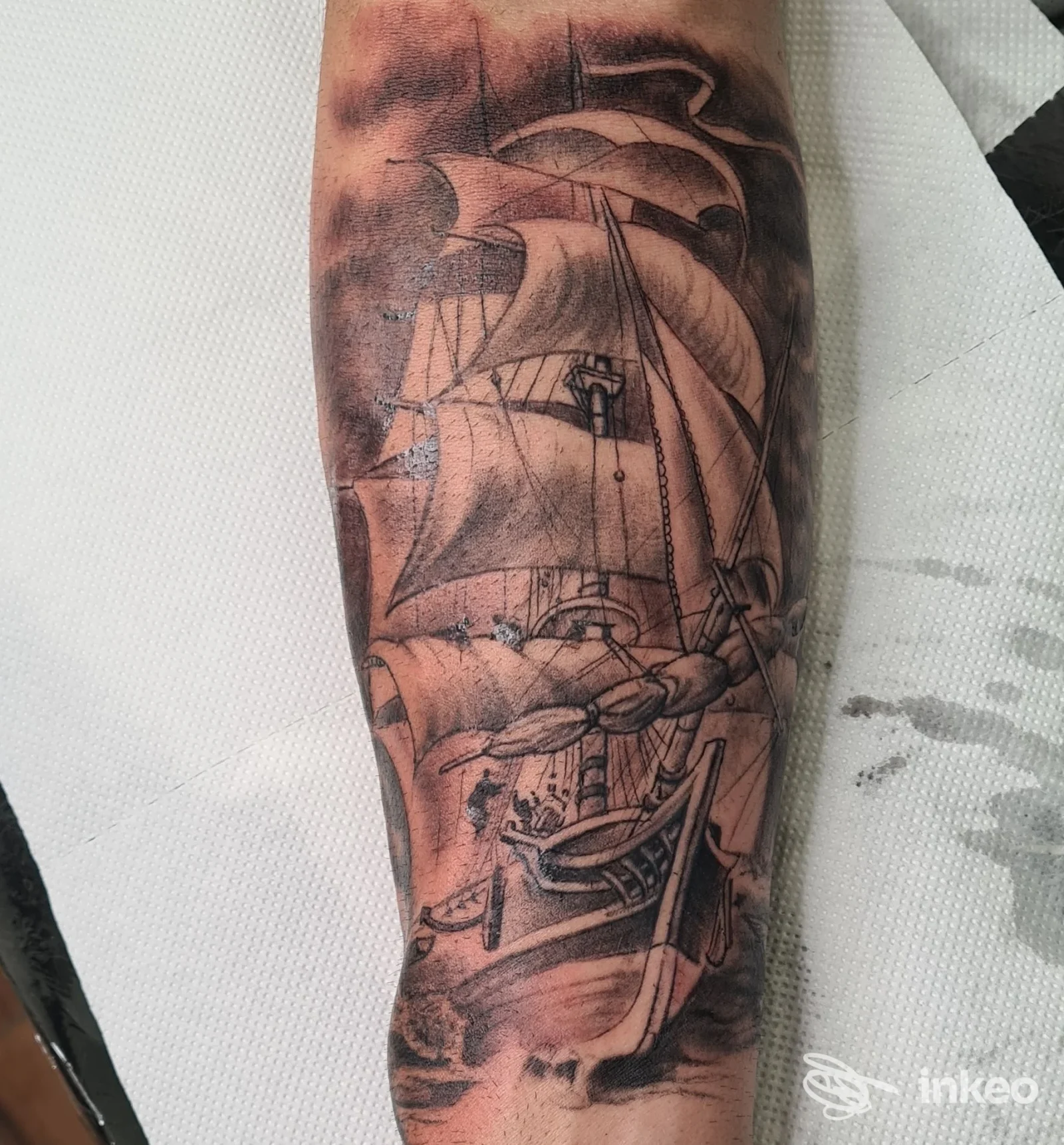Tattoo realistico di una nave su pelle, visibile sul braccio, con dettagli in bianco e nero.