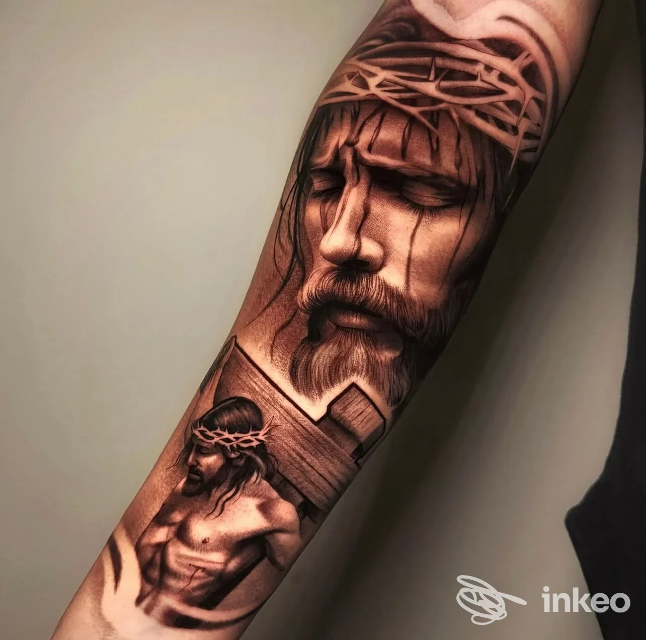 Tattoo realistico sull’avambraccio raffigurante Gesù con la corona di spine e una scena secondaria, realizzata in tonalità scure e sfumature, visibile su pelle reale.