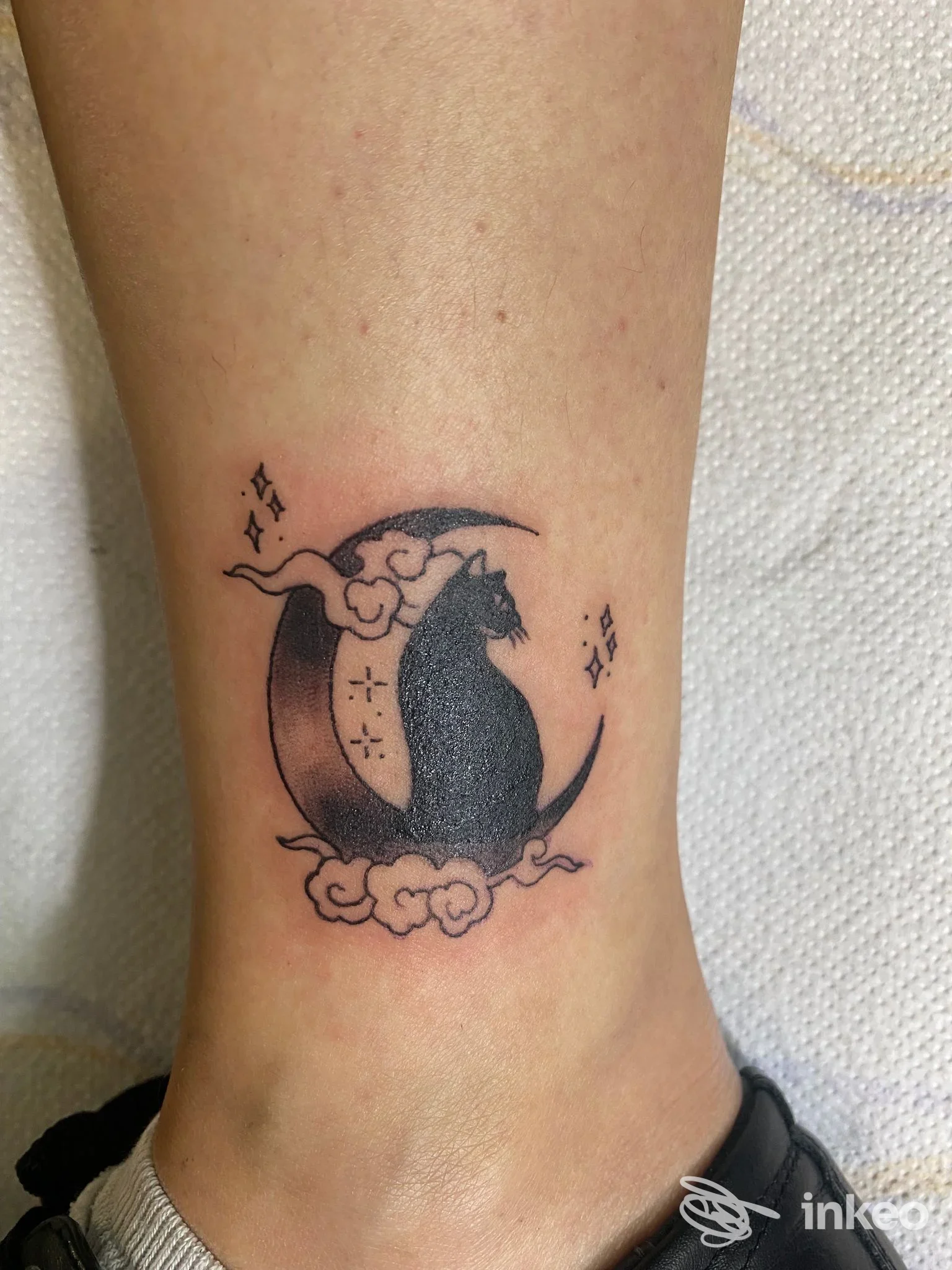 Tattoo sul polpaccio: stile grafico inchiostrato con luna, gatto nero seduto su una luna, nuvole e piccole stelle, eseguito su pelle chiara.
