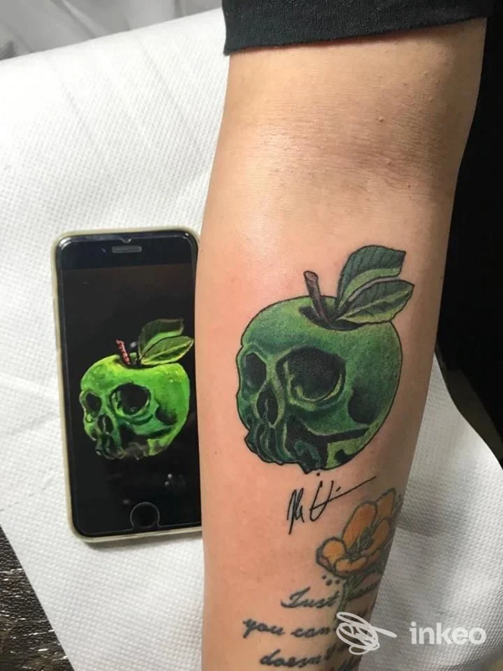 Tatuaggio sul braccio raffigurante una mela verde con teschio al centro, colorata e dettagliata; si intravede uno smartphone vicino che mostra l’immagine del tatuaggio.