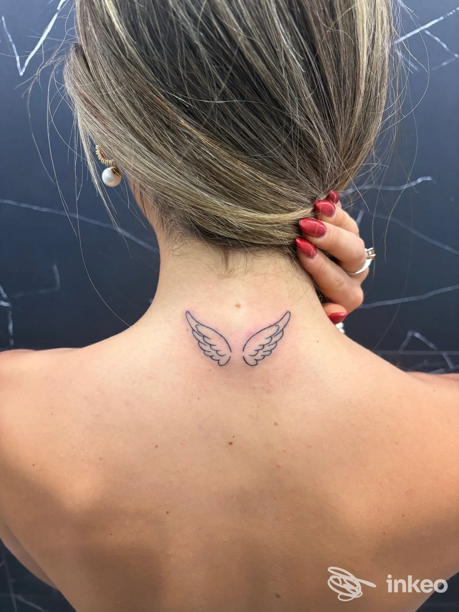 Tattoo sulle natiche della nuca: due piccole ali stilizzate in tratto nero, visibili sulla pelle della parte posteriore del collo, senza volto visibile.