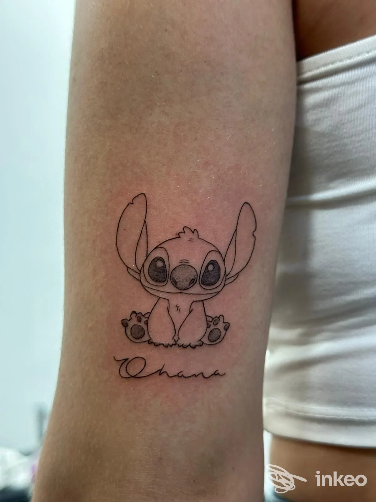 Tatuaggio sul braccio raffigurante Stitch con firma stilizzata sotto, realizzato sulla pelle con linee nette e colore limitato.
