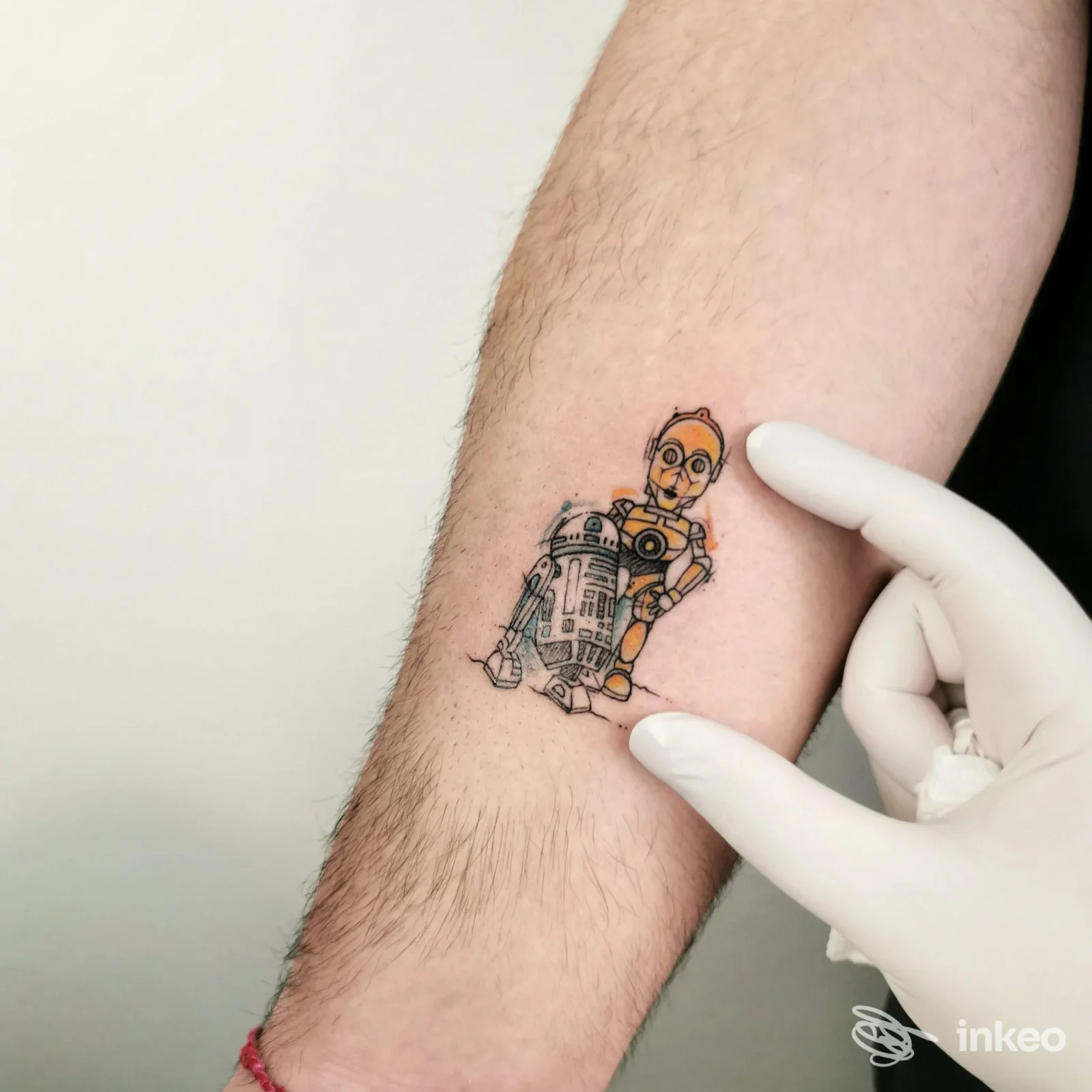 Piccolo tatuaggio su avambraccio raffigurante un robot stilizzato con contorni neri e colori giallo/arancione, mano guantata che lo mostra.