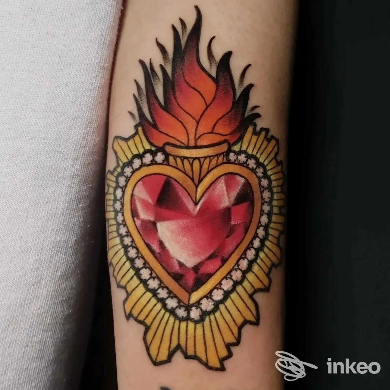 Tattoo reale su braccio: cuore al centro con fiamme rosse, contorni neri spessi e ornamenti dorati; concentrazione su stile tradizionale con dettagli bianchi come cornice.