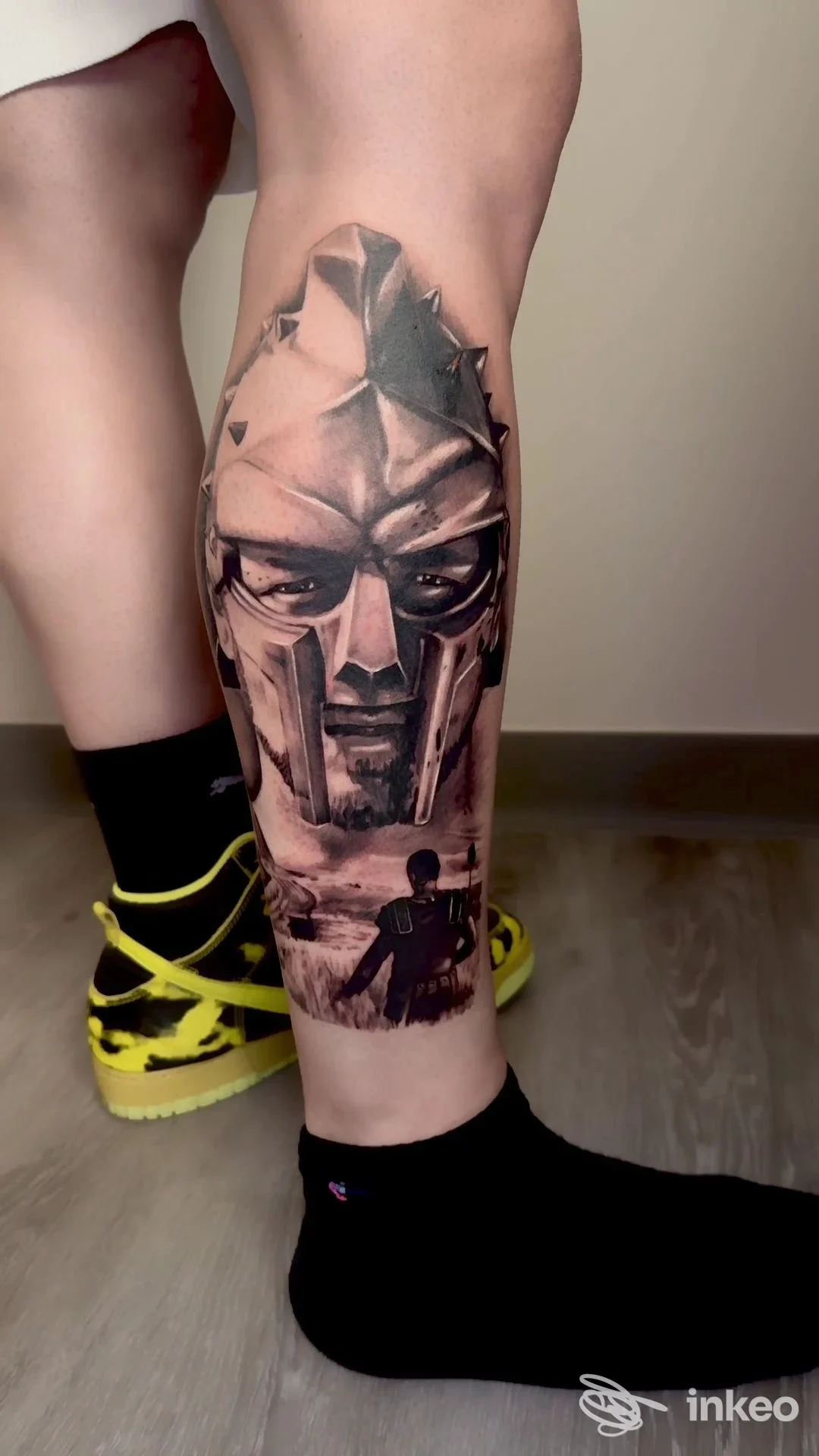 Tattoo realistico in tonalità di grigio/nero su polpaccio sinistro, raffigurante un volto maschile con elmo e figure secondarie in profondità, eseguito su pelle chiara.
