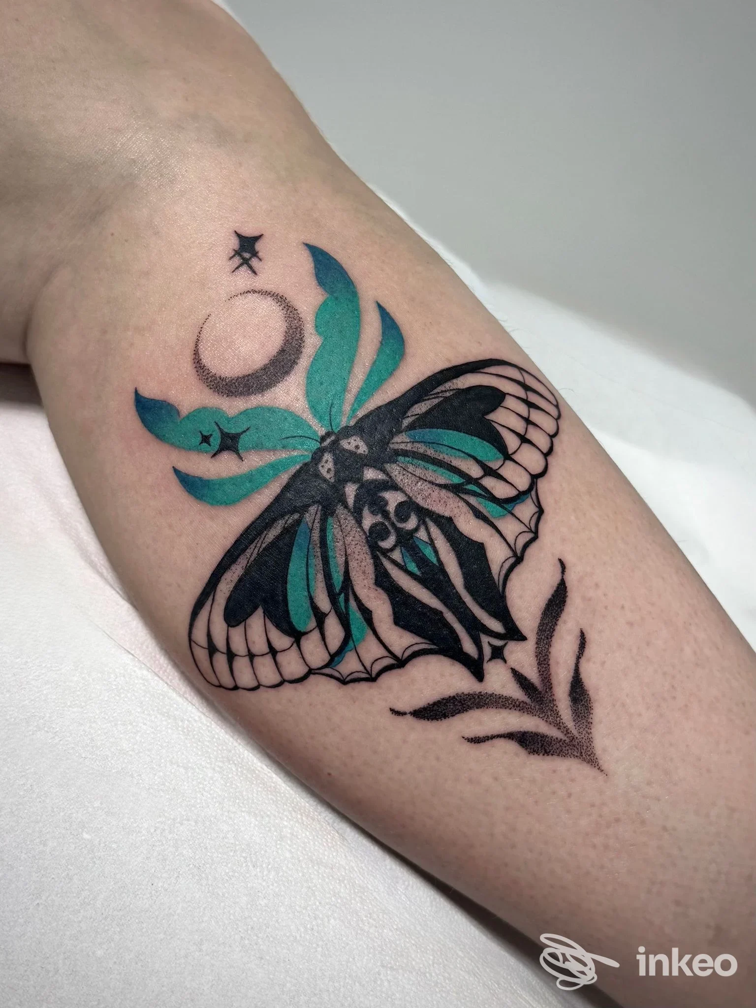 Tatuaggio su avambraccio di una farfalla stilizzata nera con ali tese, dettagli in turchese e una luna crescente, su pelle chiara.