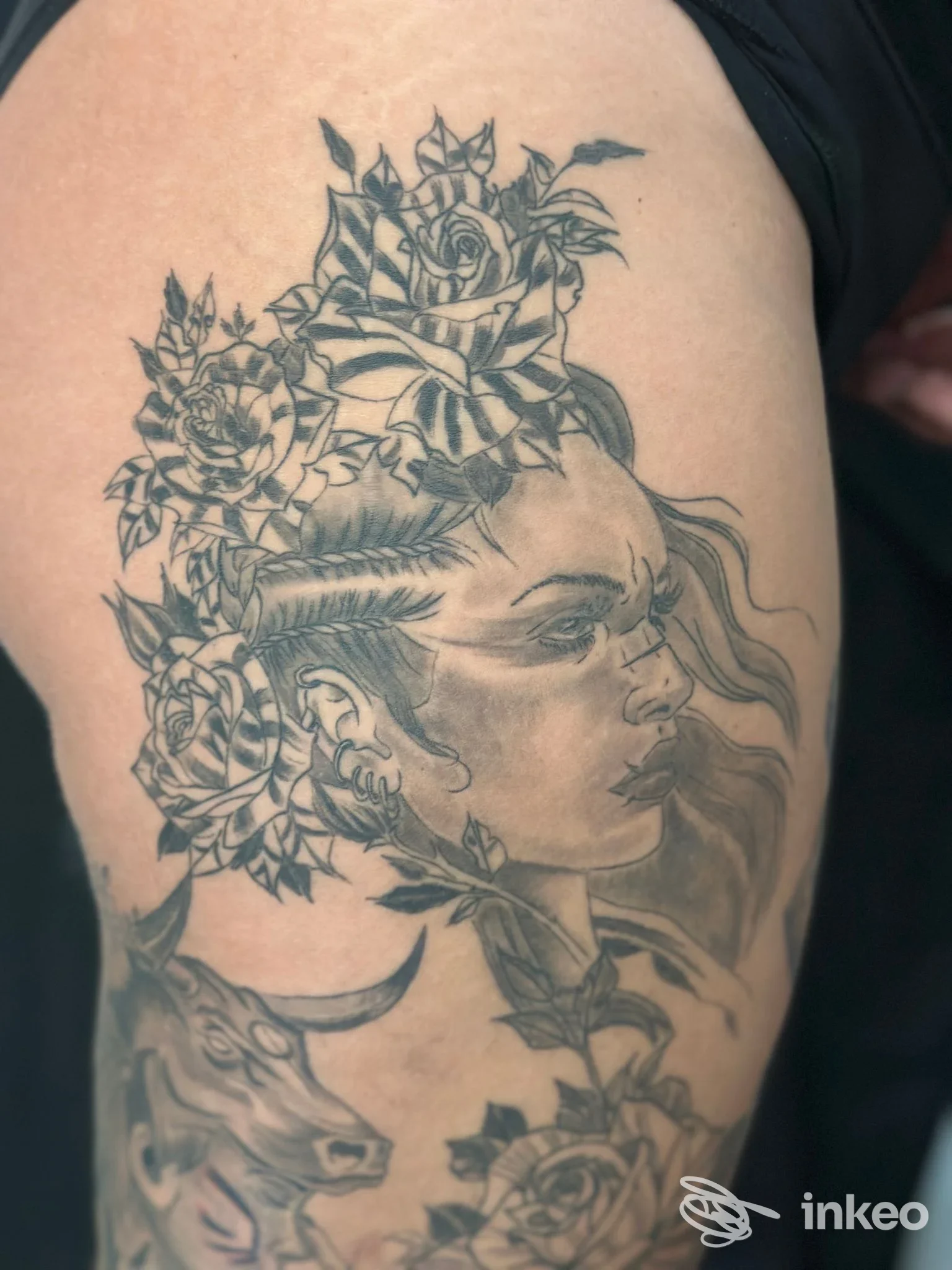 Braccio di una persona con tatuaggio ritraente una faccia femminile in profilo circondata da fiori e foglie, con una piccola figura di bestia sullo sfondo.