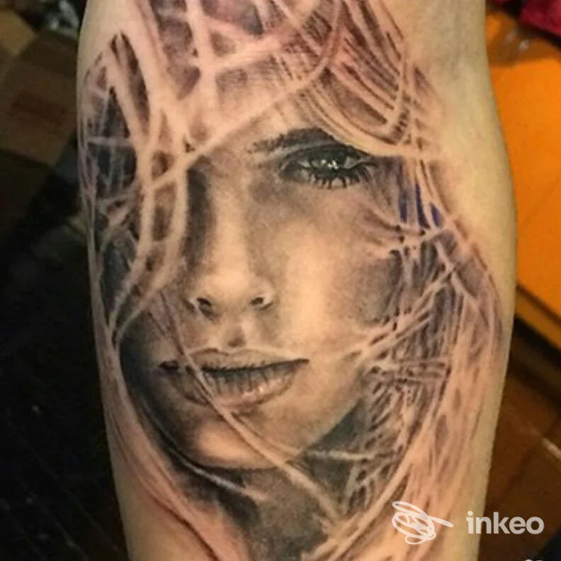 Ritratto realistico su pelle di una donna, con capelli che attraversano il viso in un tattoo ben definito, tonalità nere e grigio.