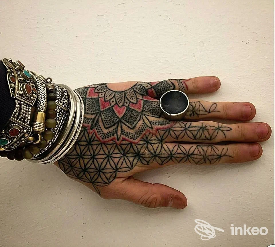 Mano su sfondo chiaro con tatuaggio geometrico a mandala sul dorso e sulle dita; bracciali e anelli decorativi al polso. Tattoo visibile su pelle reale, senza volto visibile. Riccione, INKdustria Tattoo.