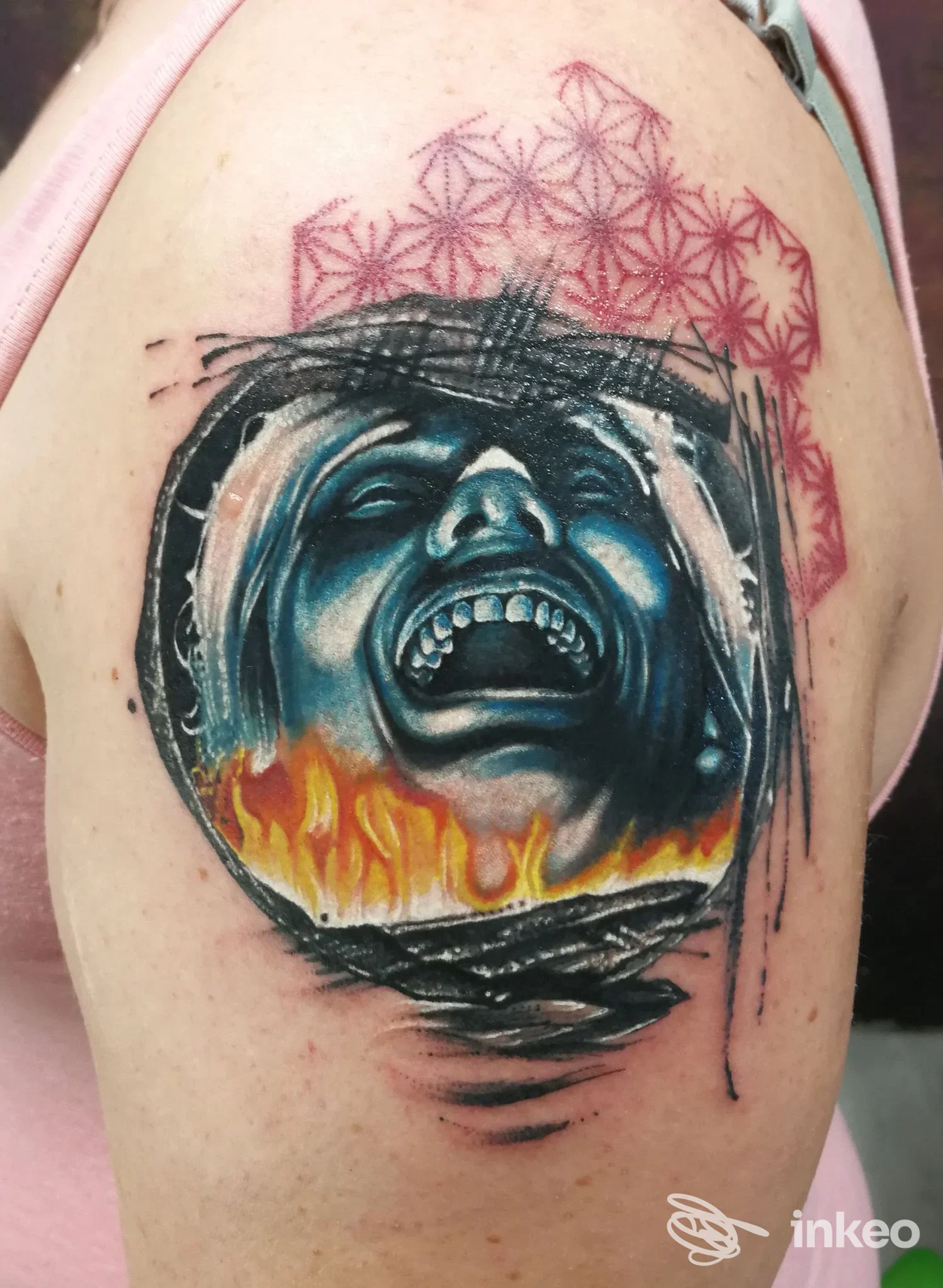Tatuaggio di una faccia urlante di colore azzurro con fiamme arancioni, contornata da motivi geometrici rosa sul braccio.