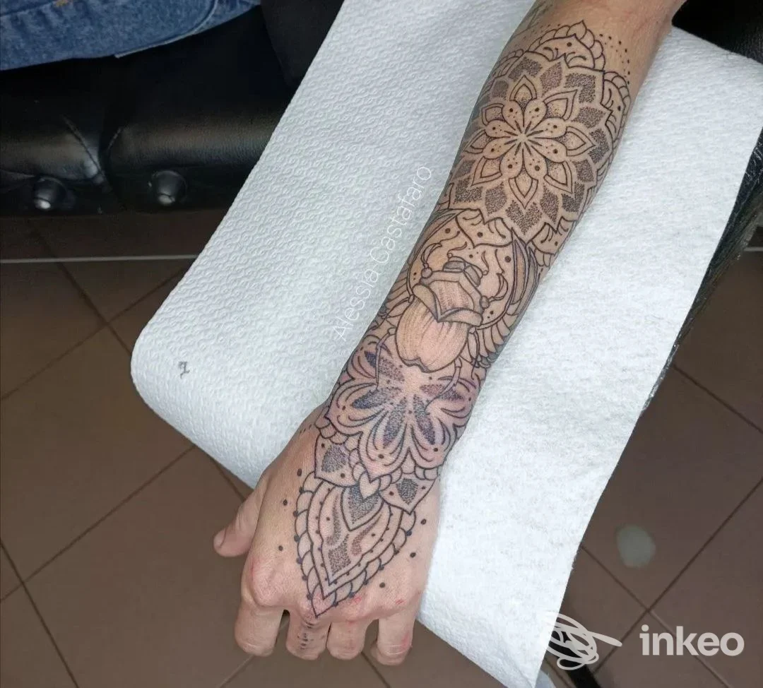 Figura il braccio sinistro con un tatuaggio esteso su avambraccio, motivi floreali geometrici, disegno inchiostrato su pelle reale, su sfondo bianco.