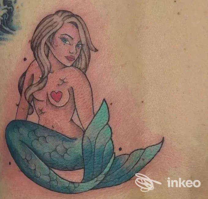 Tatuaggio colorato di una sirena su pelle reale, con coda verde-blu e cuore rosso sul petto, vista di lato sulla spalla/torace.