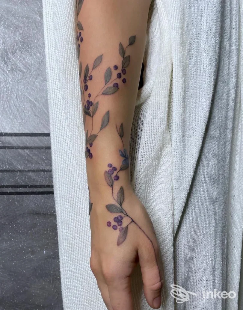 Tattoo floreale lungo l'avambraccio con piccoli frutti viola e foglie grigie, realizzato sulla pelle.