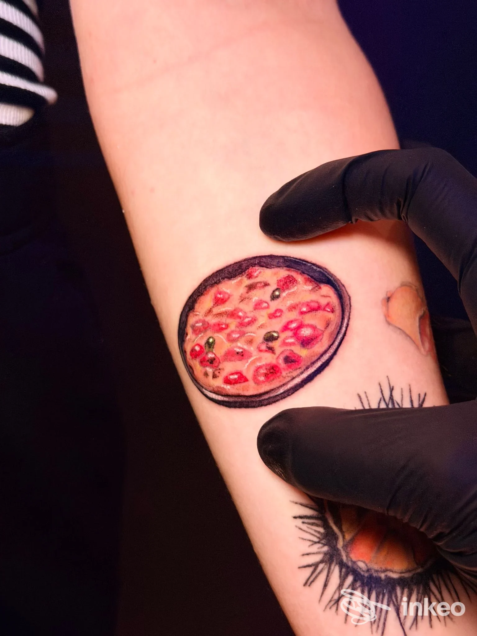 Tattoo realistico su avambraccio raffigurante una fetta di pizza, con una mano glovata che appartiene al tatuaggio e tiene la parte tatuata in primo piano.