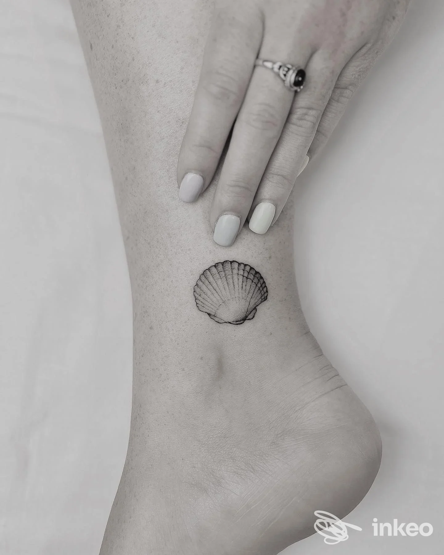 Tatuaggio a conchiglia sulla caviglia di una persona, visibile tra dita della mano e piede; stile lineare, esecuzione chiara sulla pelle.