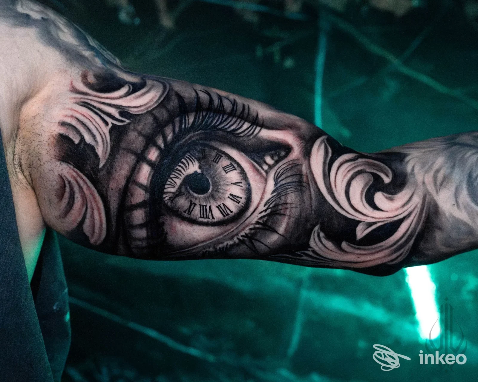 Tattoo su braccio raffigurante un occhio e un orologio, circondati da motivi decorativi scuri, con luci verdi sullo sfondo; visibile solo l'arto.